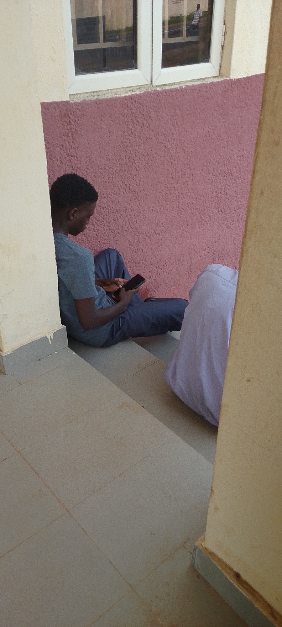In Gumel, Nigeria, a young Black male, appearing to be a teenager, is seated on a step outside, leaning against a pale yellow wall. He is wearing a dark blue t-shirt and dark blue pants, and he is holding a black smartphone in his hands, looking down at the screen. To his right, a large, light purple cloth or blanket is draped over something unseen. The background features a rough, textured pink wall with a white-framed window above it. The window reflects an outdoor scene, showing a person wearing a striped shirt. The floor is tiled in a light beige color with visible grout lines. The overall scene is outdoors, possibly in front of a building.