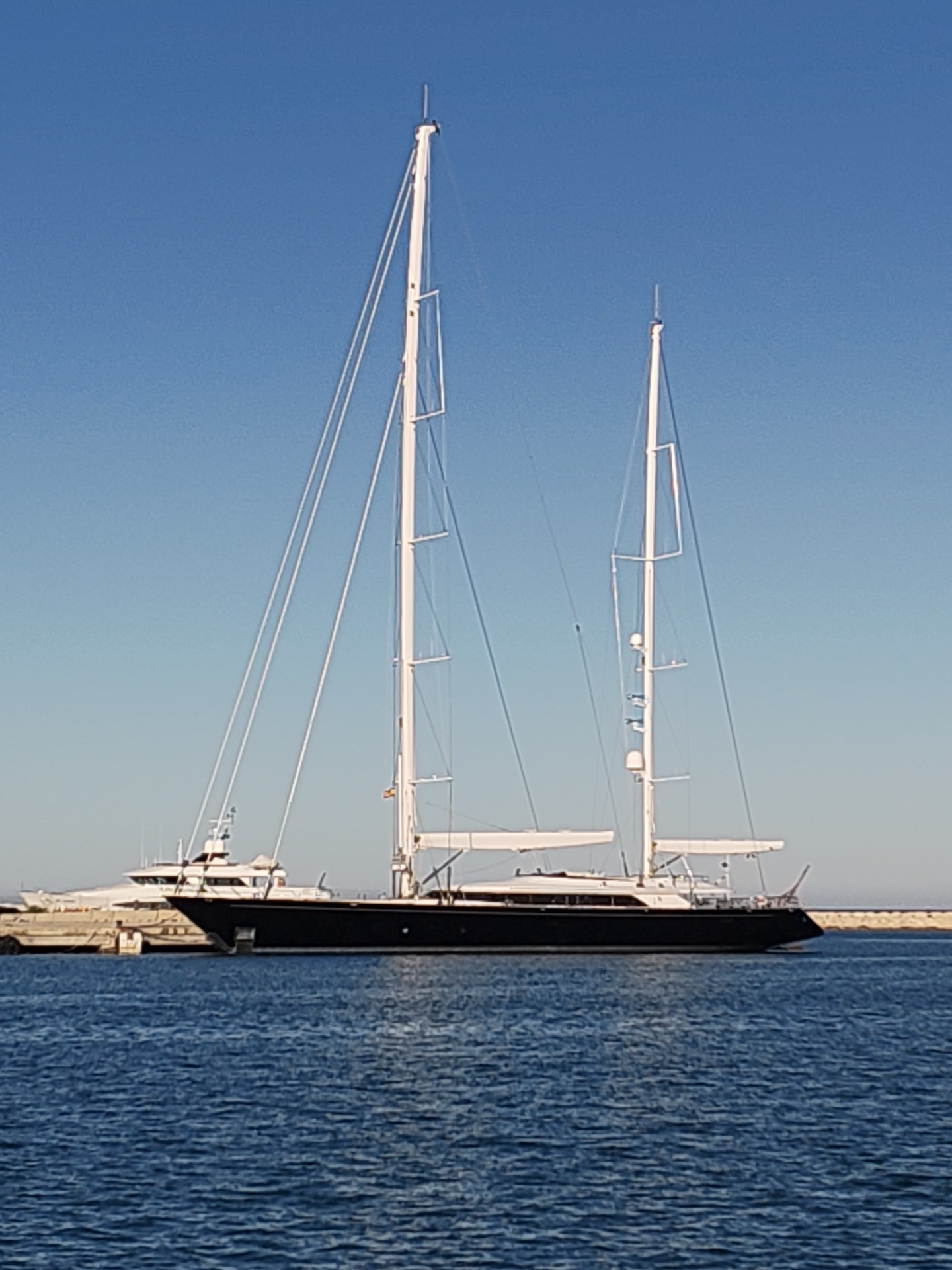 The image depicts a large, two-masted sailing yacht moored in the harbor of Dénia, Spain. The yacht has a dark hull and tall white masts with rigging. It is positioned near a breakwater, visible in the background. Another smaller yacht is also present in the harbor behind the sailing yacht. The water is dark blue and the sky is a clear, light blue. The scene appears to be during daylight with calm water conditions.