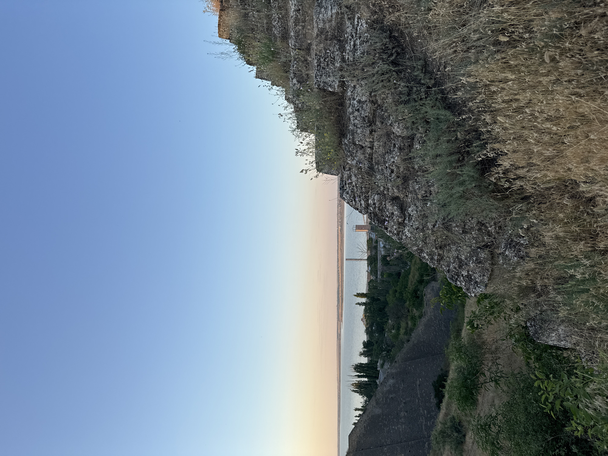 This image, taken vertically, captures a serene twilight scene overlooking a body of water and a distant coastline. The foreground is dominated by a rugged, rocky cliff face, overgrown with dry grass and sparse vegetation. The cliff is textured and appears ancient, suggesting a natural formation or an old structure.
Below the cliff, a dark, stone-built embankment or wall runs along the slope, hinting at historical fortifications or terracing. Beyond this, a vast expanse of water stretches towards the horizon. The water is calm and reflects the soft hues of the setting sun. On the far side of the water, a low-lying coastline is visible, dotted with trees and what appear to be structures, including a tall, slender tower and possibly a bridge in the distance.
The sky transitions from a clear, pale blue at the top of the frame to soft shades of pink and orange near the horizon, characteristic of sunset. The overall mood is peaceful and tranquil, evoking a sense of natural beauty and the passage of time. The lighting indicates it is likely early evening. The image was captured in Mizhlymanske, Ukraine. There is no visible text in the image.