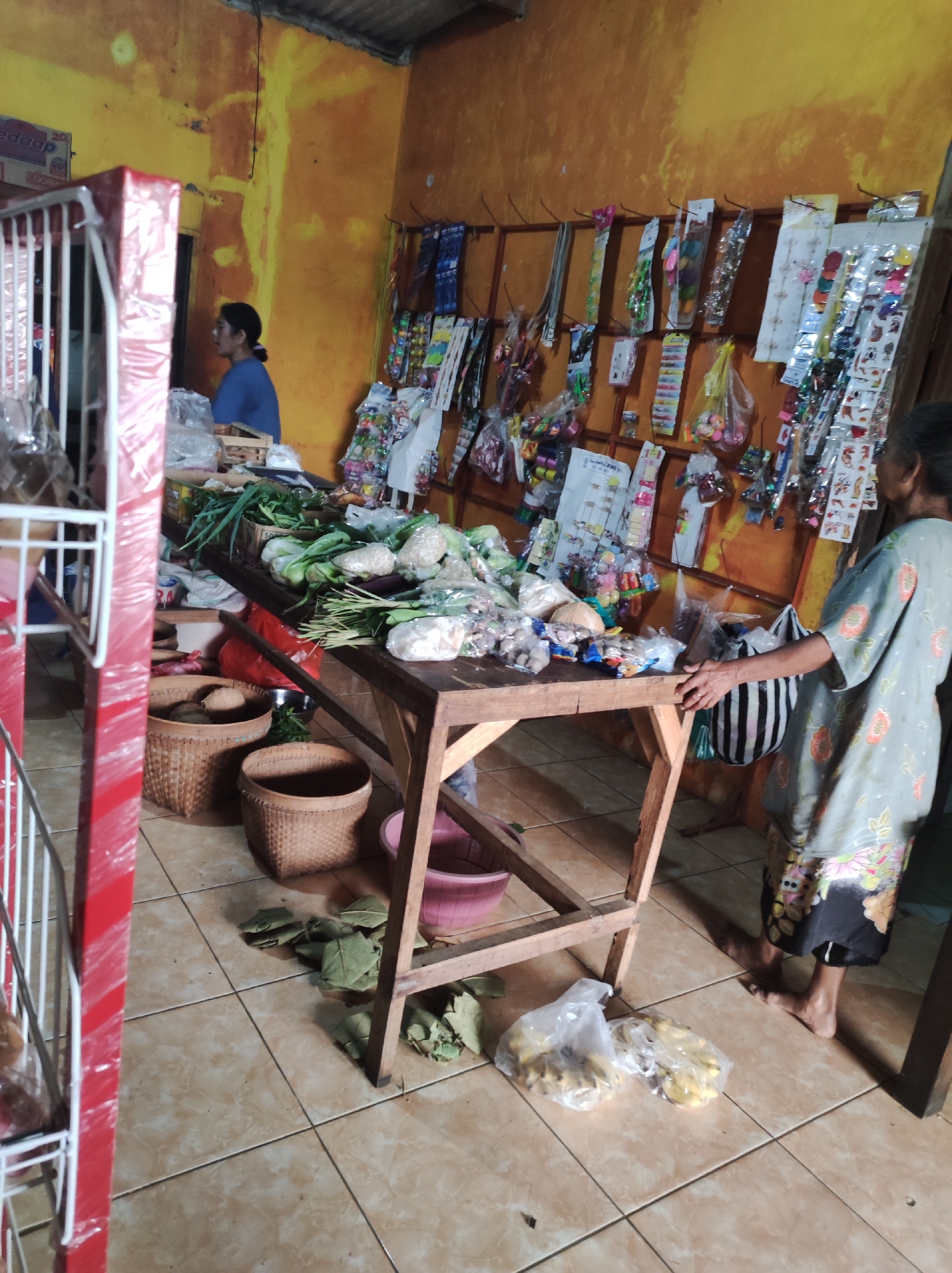 The image depicts the interior of a small shop in Mojopuro, Indonesia. The walls are painted a warm, faded yellow, with some discoloration and peeling. The ceiling appears to be corrugated metal. In the foreground, to the left, is a red metal shelving unit with white bars, partially wrapped in plastic. Adjacent to this is a wooden table displaying a variety of fresh produce, including leafy greens, vegetables like cabbage and eggplant, and what appear to be coconuts and other root vegetables. Several woven baskets are placed on the floor beneath the table, some containing items. To the right of the table, a woman in a grey patterned garment with floral accents is standing, her body angled away from the camera. She appears to be interacting with the table. On the floor near her are two plastic bags containing yellow, possibly starchy, items. Further back in the shop, another woman in a blue top is visible near a doorway. The walls are adorned with numerous items, including what seem to be small packaged goods, toys, or accessories hanging from hooks. The floor is tiled with square, beige-patterned tiles. The lighting suggests an indoor setting with natural light coming from an unseen source, creating shadows and highlighting the textures of the materials.