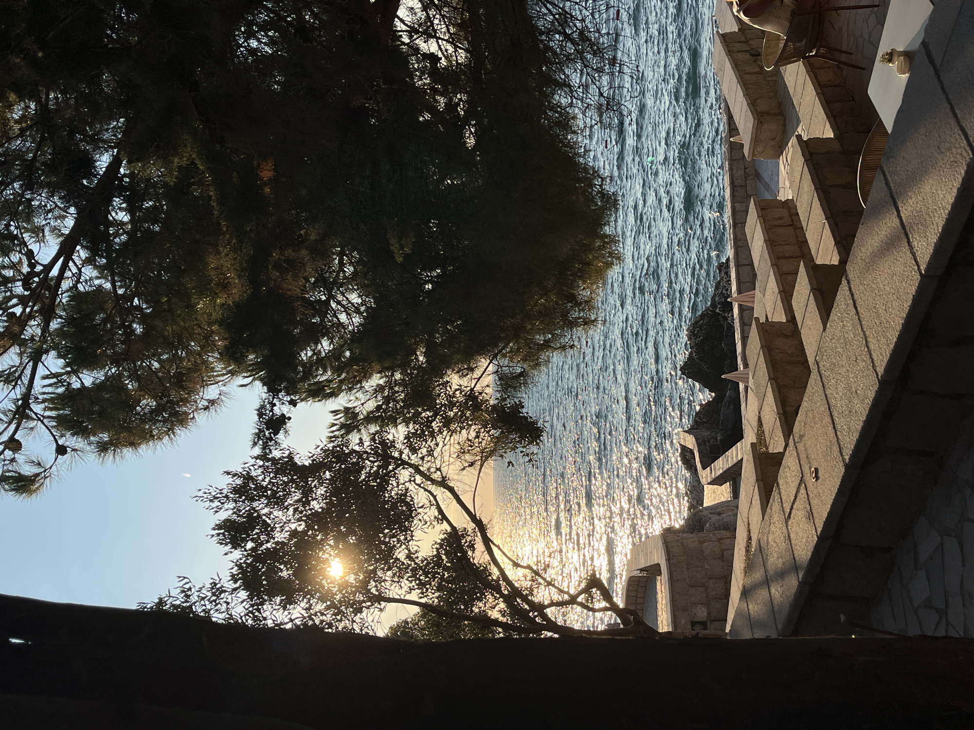 The image depicts an outdoor seating area at a seaside establishment in Pečurice, Montenegro, bathed in the warm glow of late afternoon sunlight. The foreground features a stone-paved patio with multiple levels of stone benches and tables, some with chairs. In the middle ground, a person is seated at a table, facing away from the camera, their back to the sea. Beyond the seating area, stone steps and walls lead down to the rocky coastline. The shimmering blue sea stretches to the horizon, reflecting the golden sunlight, with waves gently lapping against the shore. Lush green trees, including pine and olive trees, frame the scene from the left and right, their branches casting dappled shadows across the patio. The sky is a clear blue, with a sliver of a crescent moon visible.