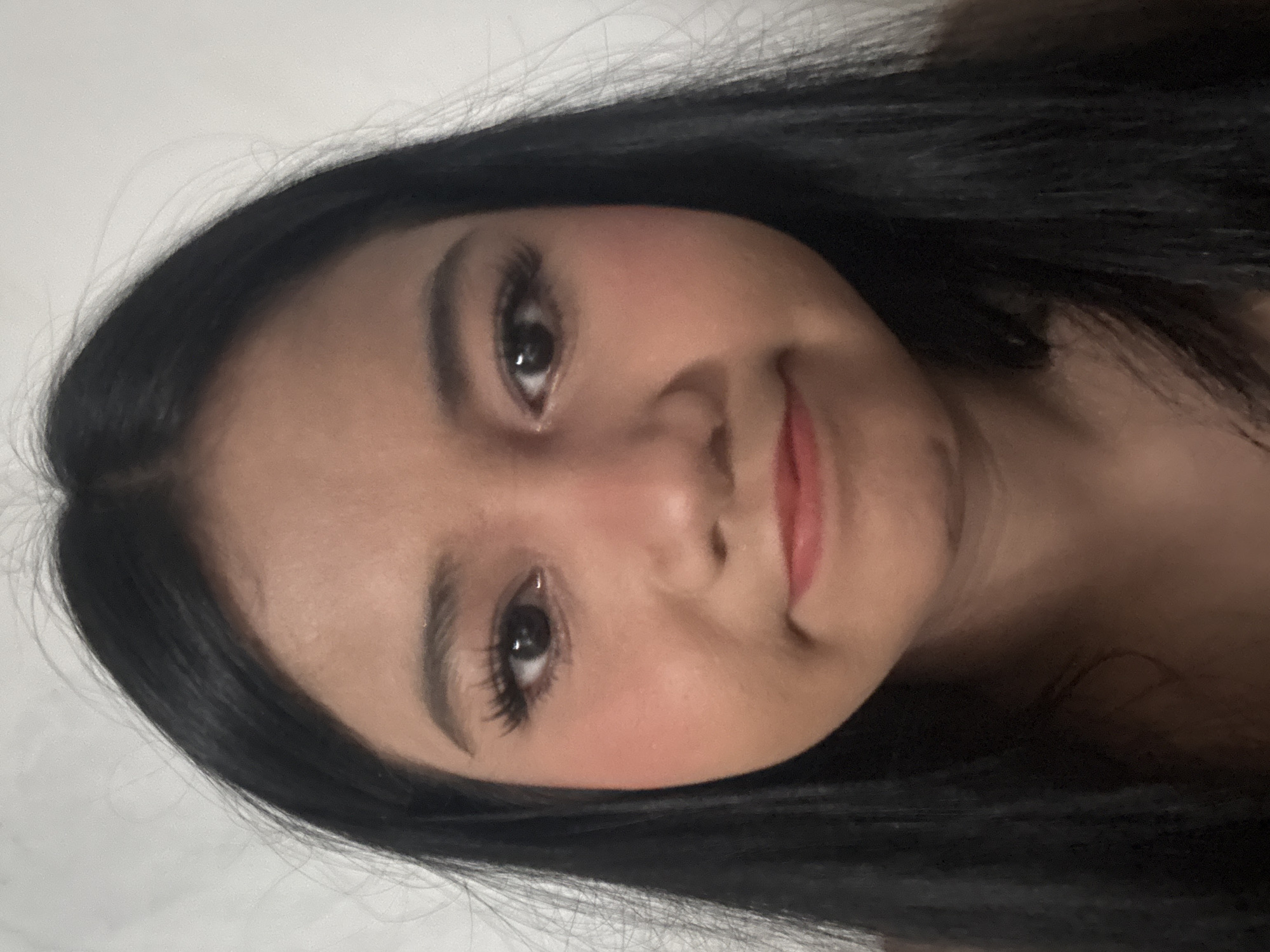A close-up, front-facing portrait of a young woman with long, dark hair frames her face. Her skin tone is light to medium, and she is wearing makeup, including blush on her cheeks, mascara, and lipstick. Her eyes are dark and open, and her mouth is slightly pursed. The background is a plain, light-colored wall. The image was taken in North Jakarta, Indonesia.