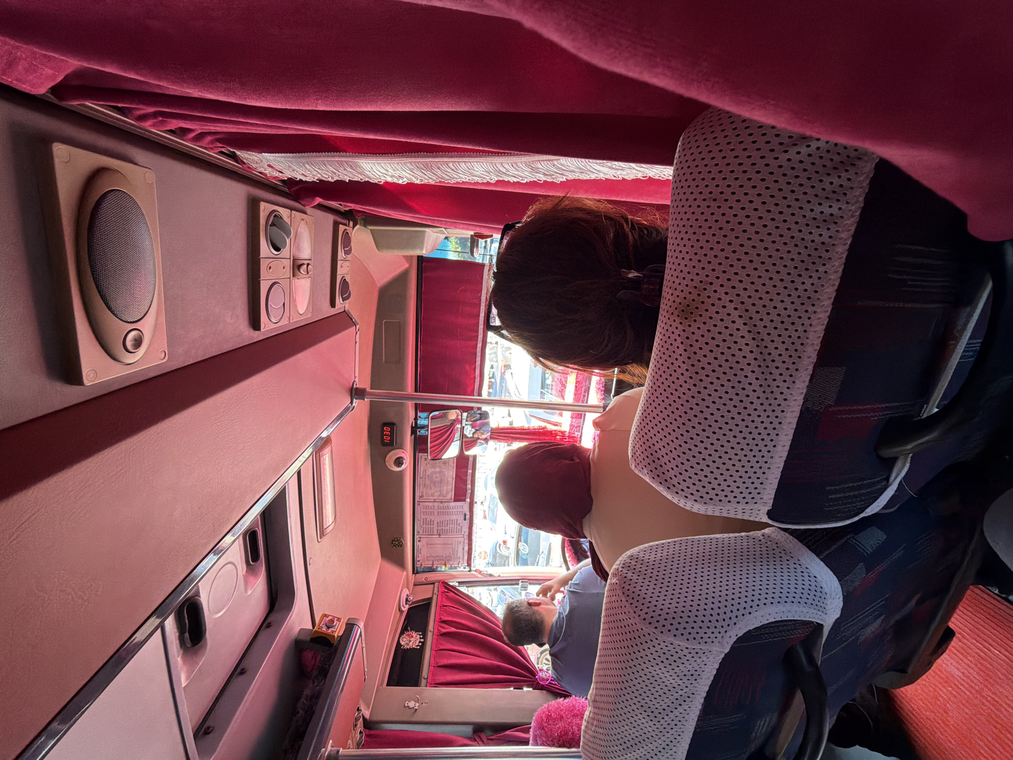 The image shows the interior of a bus, likely in Konyaaltı, Turkey, as indicated by the location context. The scene is dimly lit, with red velvet curtains covering the windows and parts of the interior, creating a somewhat luxurious but potentially claustrophobic atmosphere. The camera is positioned to capture the view from a seat, looking towards the front of the bus.
The primary subjects are the passengers. In the foreground, the head and shoulders of a woman with brown hair are visible, wearing glasses. Her posture suggests she is looking forward. Further into the bus, a man wearing a blue t-shirt is seen looking down, possibly at his phone or something in his lap. Another passenger's head, covered by a red fabric, is also visible. The seats are covered with white mesh protectors, revealing the patterned fabric of the seats underneath.
Notable objects include speakers mounted on the wall, control panels with buttons and switches, and a digital clock displaying "1030". There are also mirrors reflecting the interior and exterior. Red cushions add a splash of color to the otherwise muted tones.
The overall impression is that of a typical bus journey. The time of day is not explicitly stated, but the lighting suggests it could be daytime, possibly late morning or afternoon, given the bright light filtering through the windows. There are no obvious cues about the weather, but the bright light suggests it is not raining. The emotion conveyed is difficult to ascertain, but the passengers appear to be relaxed or occupied with their own activities during the journey. The red curtains and upholstery might suggest a slightly older or more traditional bus design. The digital clock indicates a time of 10:30, but it's unclear if this refers to AM or PM, or if it's the actual time or a setting.