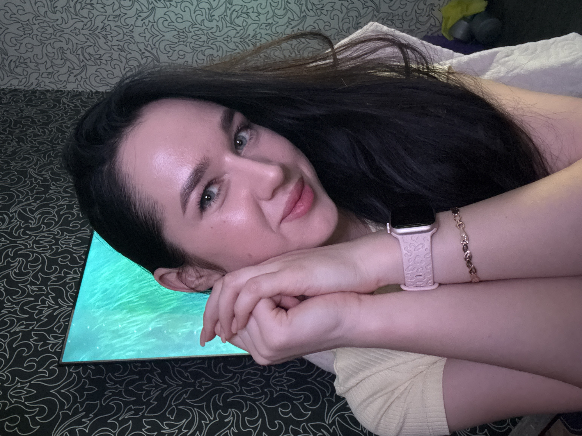 A young woman with dark hair and blue eyes is pictured from the chest up, looking at the camera with a slight smile. Her hands are clasped together in front of her face. She is wearing a light yellow shirt, a pink smartwatch, and a delicate bracelet. The background consists of dark patterned wallpaper and a large screen displaying a bright green image. The scene appears to be indoors, likely in a residential setting. There is no identifiable information in the image that would indicate its location in Novosibirsk, Russia.