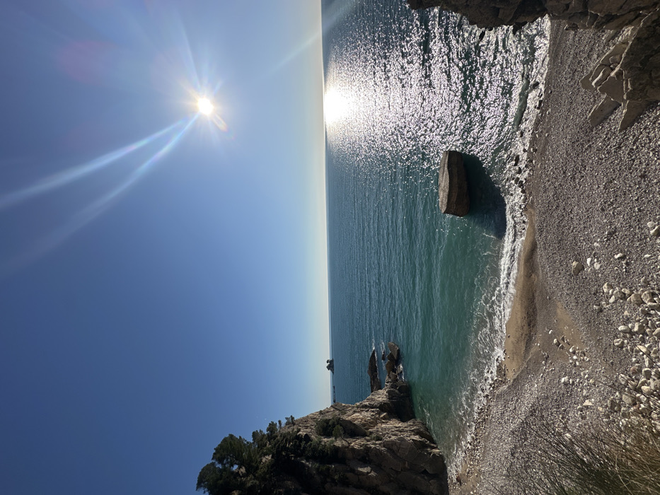 The image depicts a coastal scene at Perazića Do, Montenegro, under a clear, bright blue sky. The sun is positioned in the upper right quadrant, emitting a distinct sunburst and lens flare. The horizon separates the sky from the expansive, calm Adriatic Sea, which ranges in color from light teal near the shore to a deeper blue further out. A prominent reflection of the sun creates a shimmering path across the water extending towards the viewer.
On the left, a steep, rocky cliff face, primarily grey and brown, rises vertically, topped with a dense growth of green trees and vegetation. At the base of this cliff, smaller rocks are visible at the water's edge. In the distant sea, to the left of the sun's reflection, a small, dark islet or rock formation is discernible.
In the foreground and mid-ground, a crescent-shaped pebble beach is visible, composed of numerous light grey and brown stones, with a small section of darker sediment where the gentle waves break. A notable feature on the right side of the beach is a large, rectangular, dark-colored rock or concrete block partially submerged in the water near the shore. No individuals or man-made structures other than the aforementioned block are present within the immediate scene.