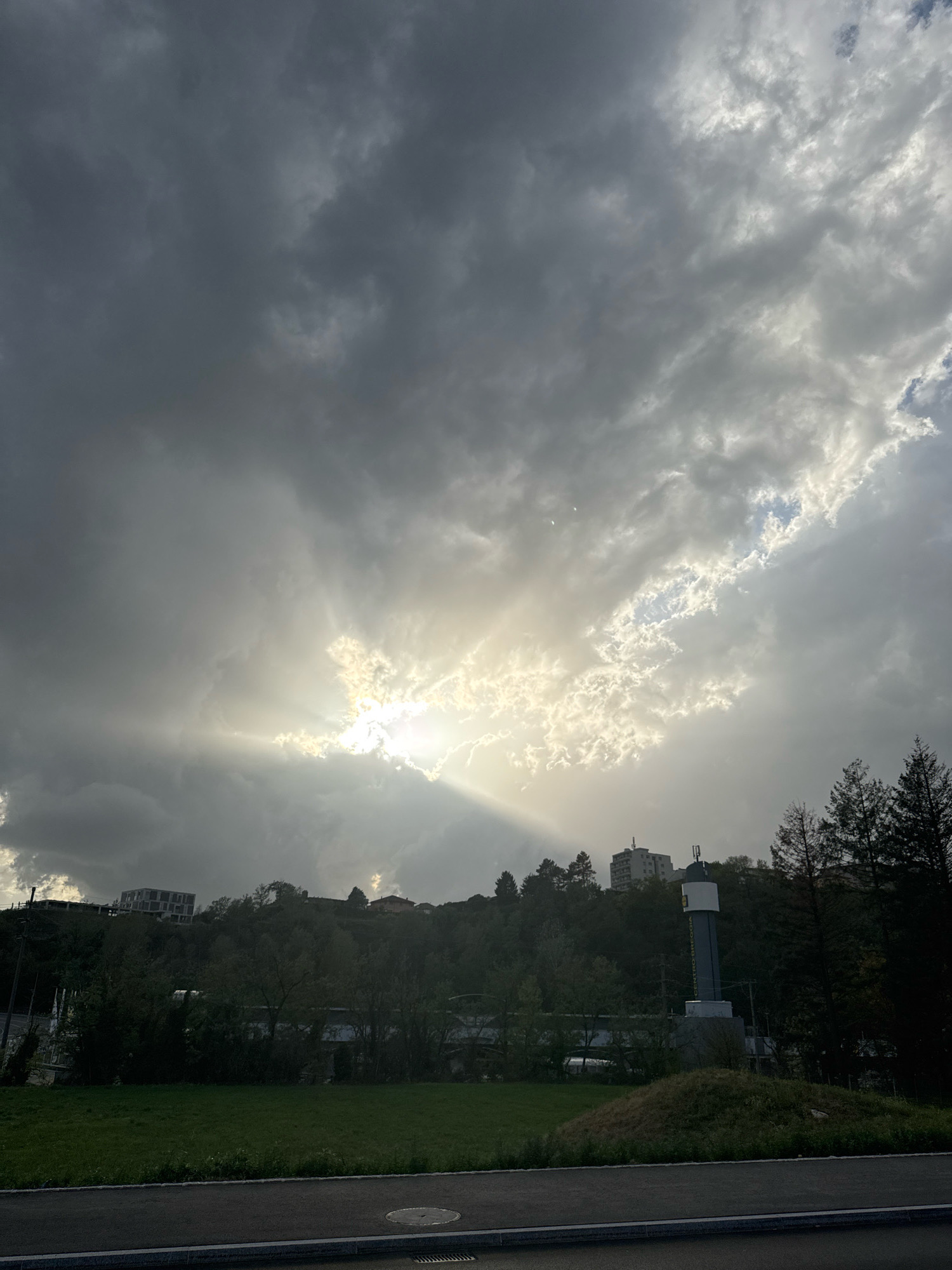 The image shows a view of the town of Morbio Inferiore, Switzerland, under a dramatic sky. In the upper portion of the frame, thick, dark clouds are interspersed with rays of sunlight breaking through, creating a dramatic, almost ethereal effect. Below the sky, a densely wooded hillside dominates the mid-ground. On top of the hill, buildings are visible, including a modern, multi-story structure on the left and several smaller houses. A distinctive tall, cylindrical water tower with a white top and a dark band featuring text is prominently positioned on the right side of the hillside, surrounded by dark green coniferous trees. In the foreground, a strip of green grass leads to a paved area, likely a road or sidewalk, with a circular manhole cover visible. The overall scene suggests a late afternoon or early evening setting with impending or passing rain. There are no people or animals visible in the frame.