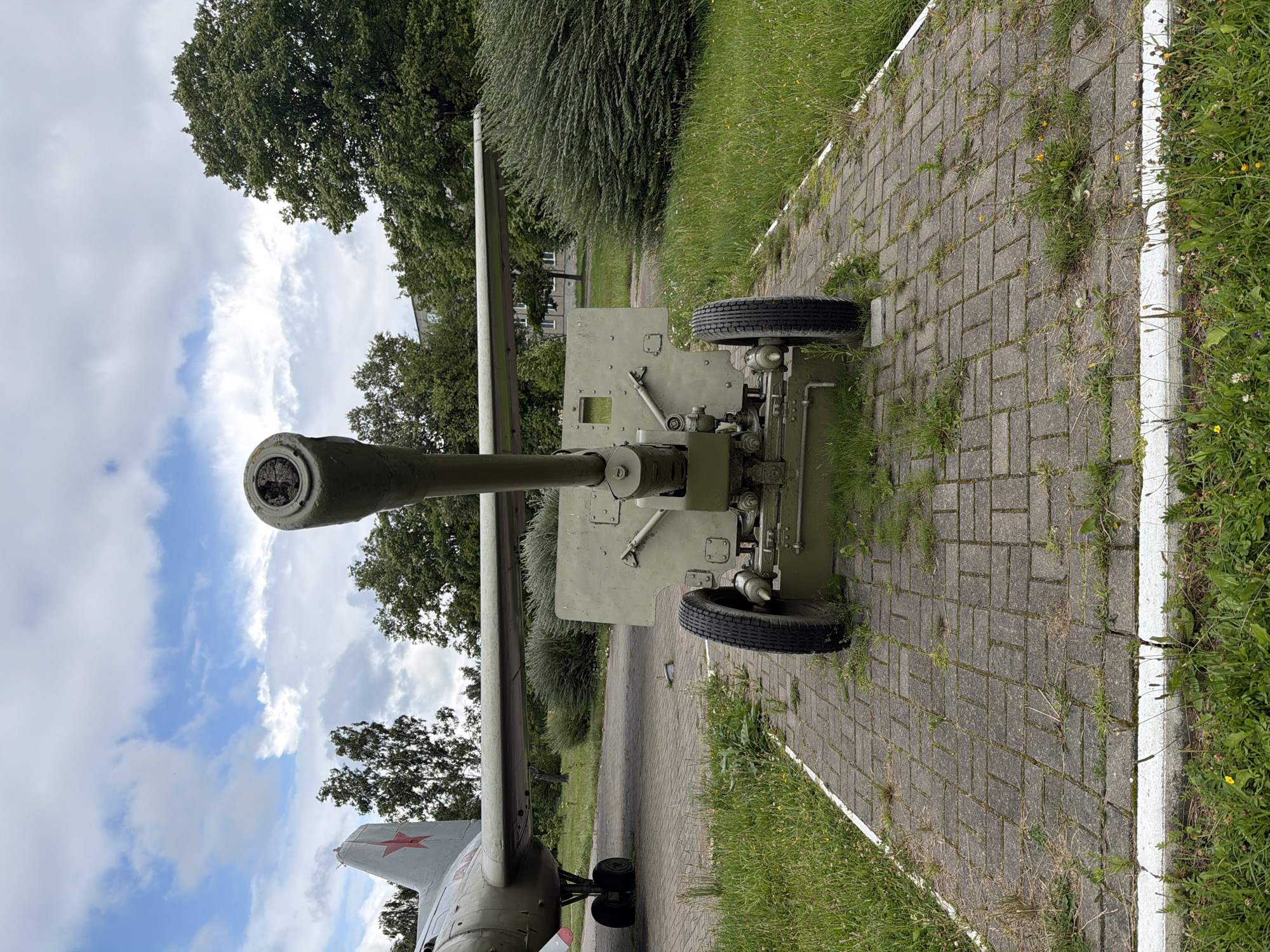 In Бягомльскі сельскі Савет, Belarus, a pale green artillery piece, possibly a field gun, is displayed on a paved walkway. The gun is positioned front and center, with its barrel pointing forward and slightly upwards. It is mounted on a wheeled carriage with two large tires. Behind the artillery piece, the wing and tail of a grey aircraft, bearing a red star insignia, are visible. The aircraft is parked on a paved surface. The scene is outdoors, with lush green grass and trees forming the background. The sky is partly cloudy. The setting appears to be an outdoor exhibition or memorial site.