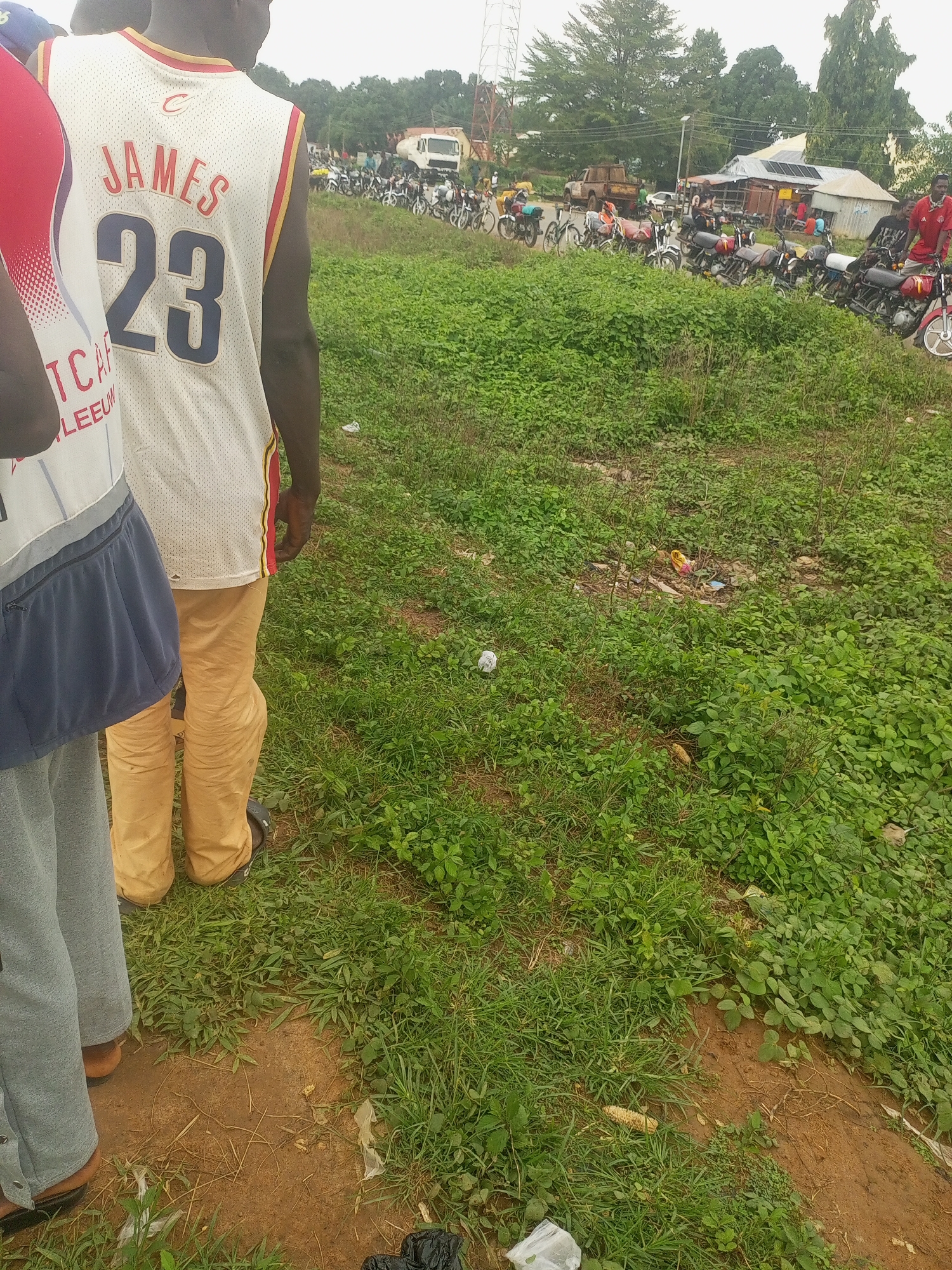 In Serti, Nigeria, a group of individuals stands near a street lined with parked motorcycles. One person in the foreground wears a white basketball jersey with the name "JAMES" and the number "23" prominently displayed on the back. This individual is also wearing tan pants. Adjacent to them, another person is visible, wearing a gray tracksuit. The background shows a street scene with buildings, a white truck, and a substantial number of motorcycles parked along the roadside, indicating a gathering or marketplace. The environment is characterized by overgrown green vegetation bordering the road, with some visible litter scattered in the grass. The sky appears overcast.