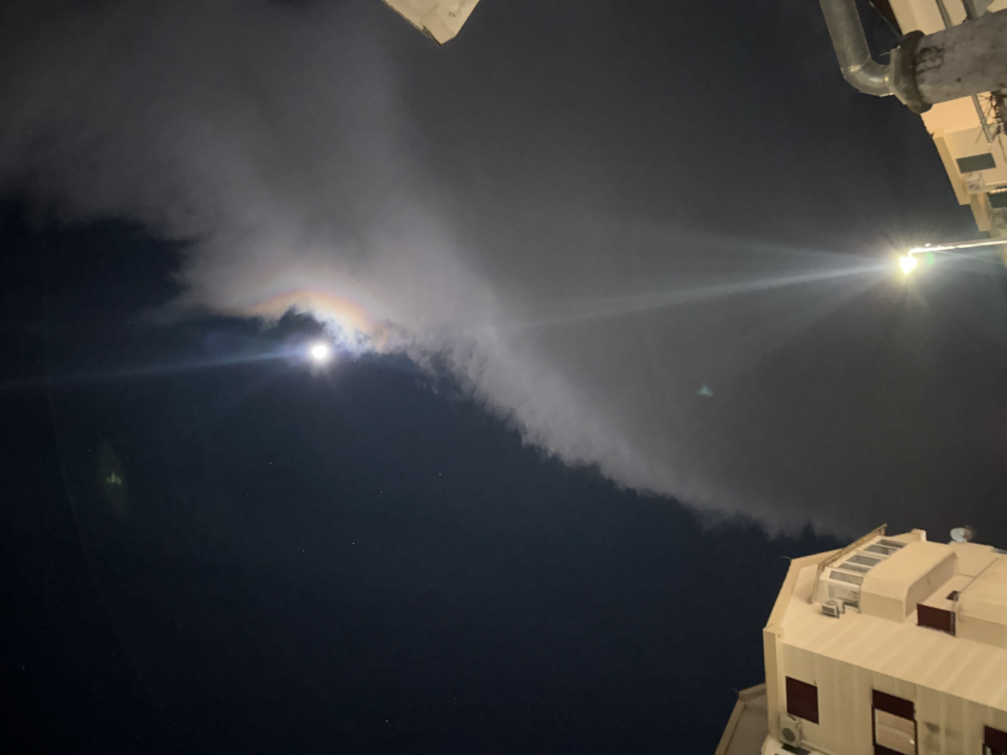The image depicts a night scene in an urban environment. A wispy cloud stretches across the sky, illuminated by the moon, which is visible through the cloud, creating a rainbow effect. A street light shines brightly in the upper right. The lower part of the image includes the rooftops of several buildings. The city cannot be identified from this image.