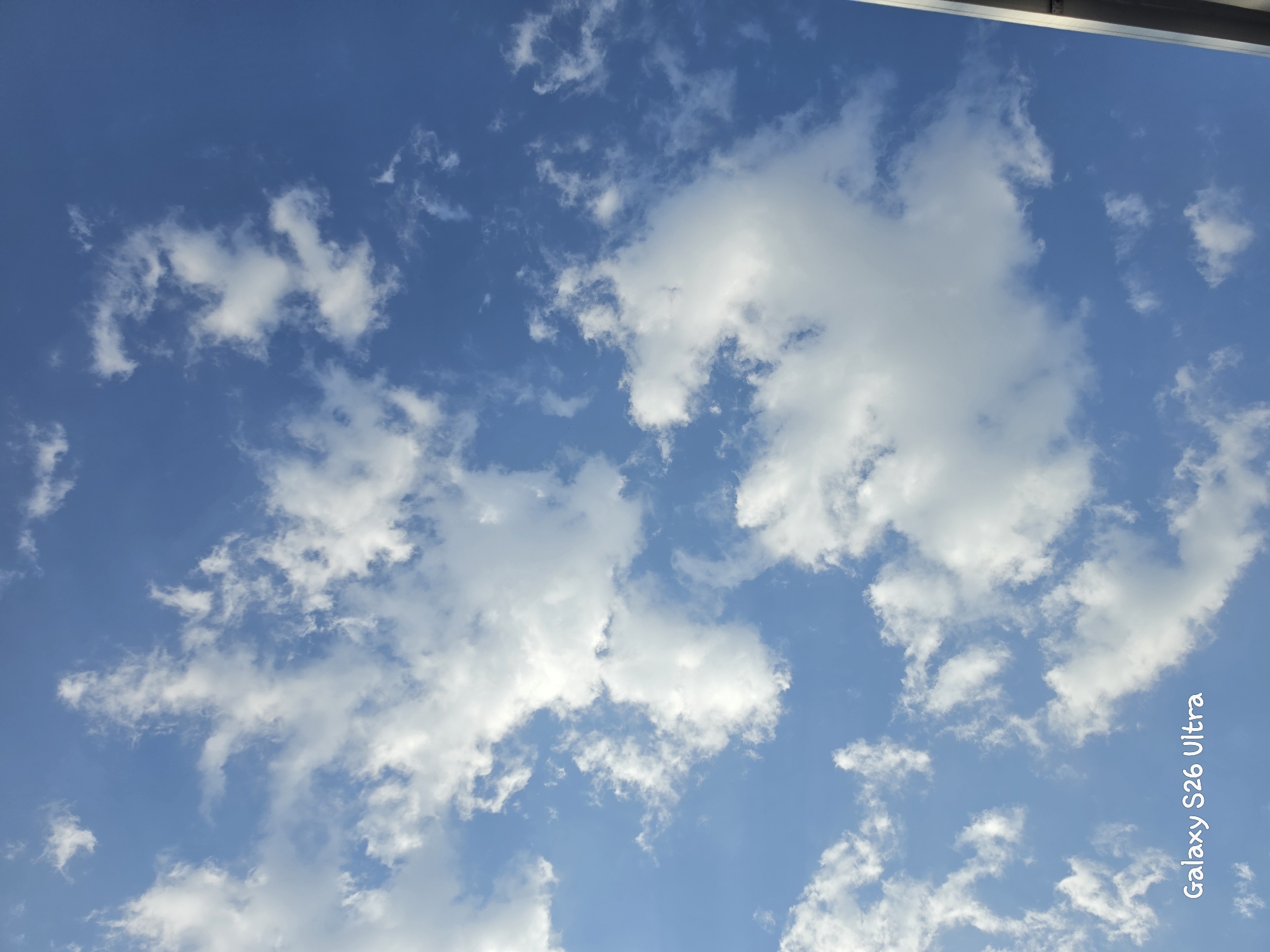 The media file captures a sky filled with white, puffy clouds against a clear blue backdrop. The clouds vary in size and density, with some appearing wispy and scattered, while others are more substantial and textured. The overall impression is of a bright, possibly sunny day. The image is oriented vertically, suggesting it might have been taken looking upwards.
In the lower right corner, vertically oriented text is visible. It reads "Galaxy S26 Ultra" in a stylized white font. This indicates the image was likely captured with a Samsung Galaxy S26 Ultra smartphone.
Without further context, it is difficult to determine the specific activity or event taking place. However, the scene suggests a moment of observation of the sky, perhaps during a casual outing or while enjoying the outdoors. The weather appears pleasant, with the presence of clouds suggesting dynamic atmospheric conditions. The location context provided is Plovdiv, Bulgaria, but this cannot be visually confirmed from the image itself. The time of day is implied to be daytime due to the brightness of the sky and the color of the clouds. No emotions are explicitly conveyed, but the scene could evoke feelings of peace, wonder, or contemplation of nature.