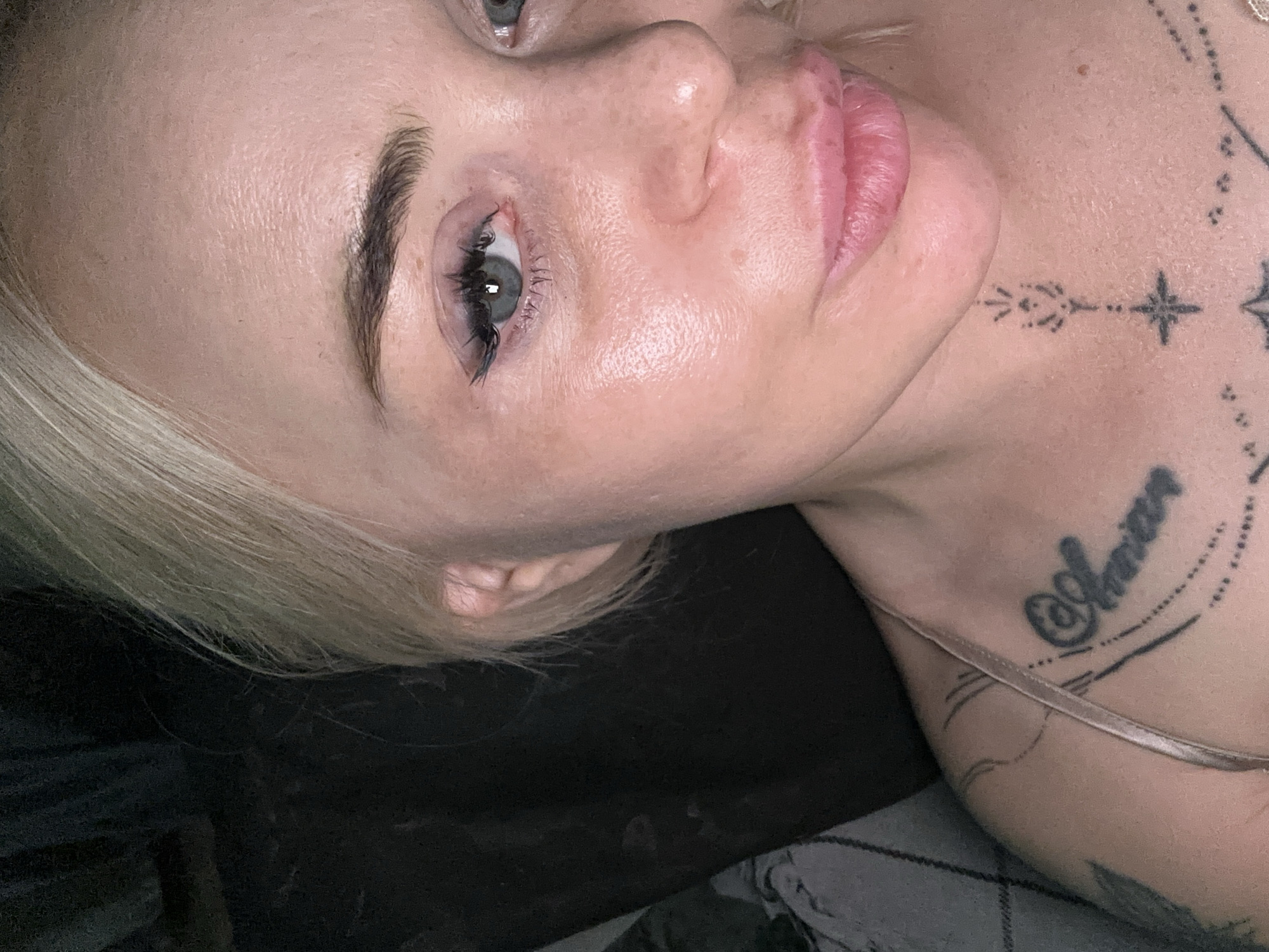 A close-up, eye-level portrait shot captures an adult individual, appearing female, with short blonde hair styled to the left, partially obscuring the left side of her face and ear. Her light-colored eyes, likely blue or grey, are open and directed straight at the camera. Her right eye (viewer's left) displays dark eyeliner and mascara on the upper lash line. Her skin is fair, exhibiting a natural texture with some areas of visible pores and a slight sheen, particularly on her cheeks and forehead. Her full, pink lips are slightly parted, forming a subtle, closed-mouth smile.
She is wearing a thin, light-colored strap from a garment over her left shoulder. Multiple tattoos are visible: on her right upper chest/clavicle area, a stylized text tattoo, possibly reading "@Aman," is present. On her lower neck/sternum, a vertical geometric tattoo with a star at its base extends upwards. Faint, curved lines of another tattoo are visible on her left shoulder/upper arm.
The background is dark and largely indistinct, suggesting an indoor environment with low light. To the viewer's left, a dark grey patterned fabric, possibly a blanket or cushion with a subtle checkered design, is partially visible. The lighting appears to be primarily from the front, illuminating the subject's face. The location is unknown.