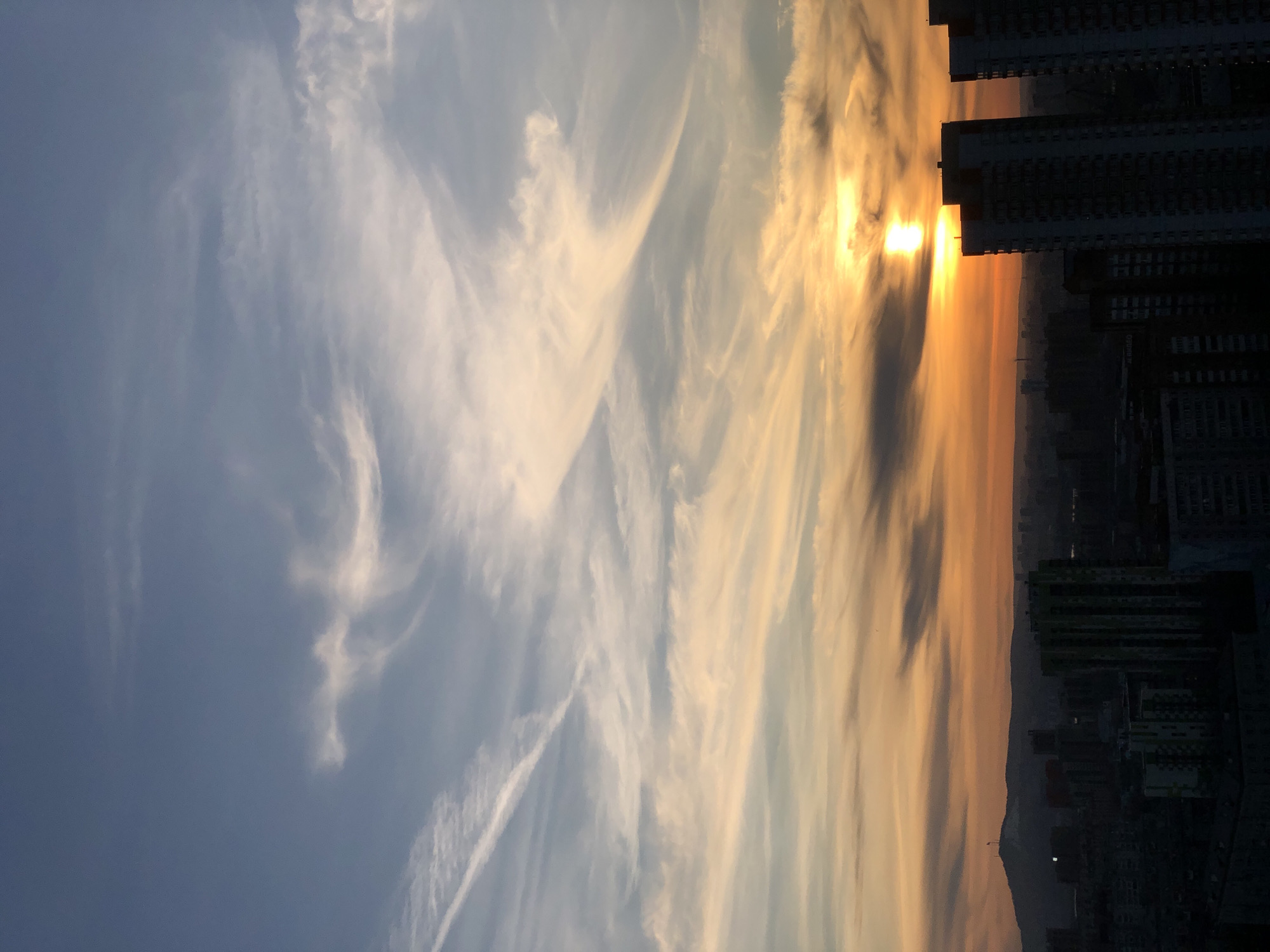 Here's a description of the image:
The image shows a sunrise or sunset in a city, likely Krasnoyarsk, Russia. The sky is filled with cirrus clouds, some illuminated with a golden hue from the sun. The sun is positioned low on the horizon, partially obscured by the silhouettes of tall buildings. These buildings dominate the right side of the frame, their structures appearing as dark blocks. In the distance, there's a hint of a cityscape and hills. The overall atmosphere is one of a serene urban dawn or dusk.