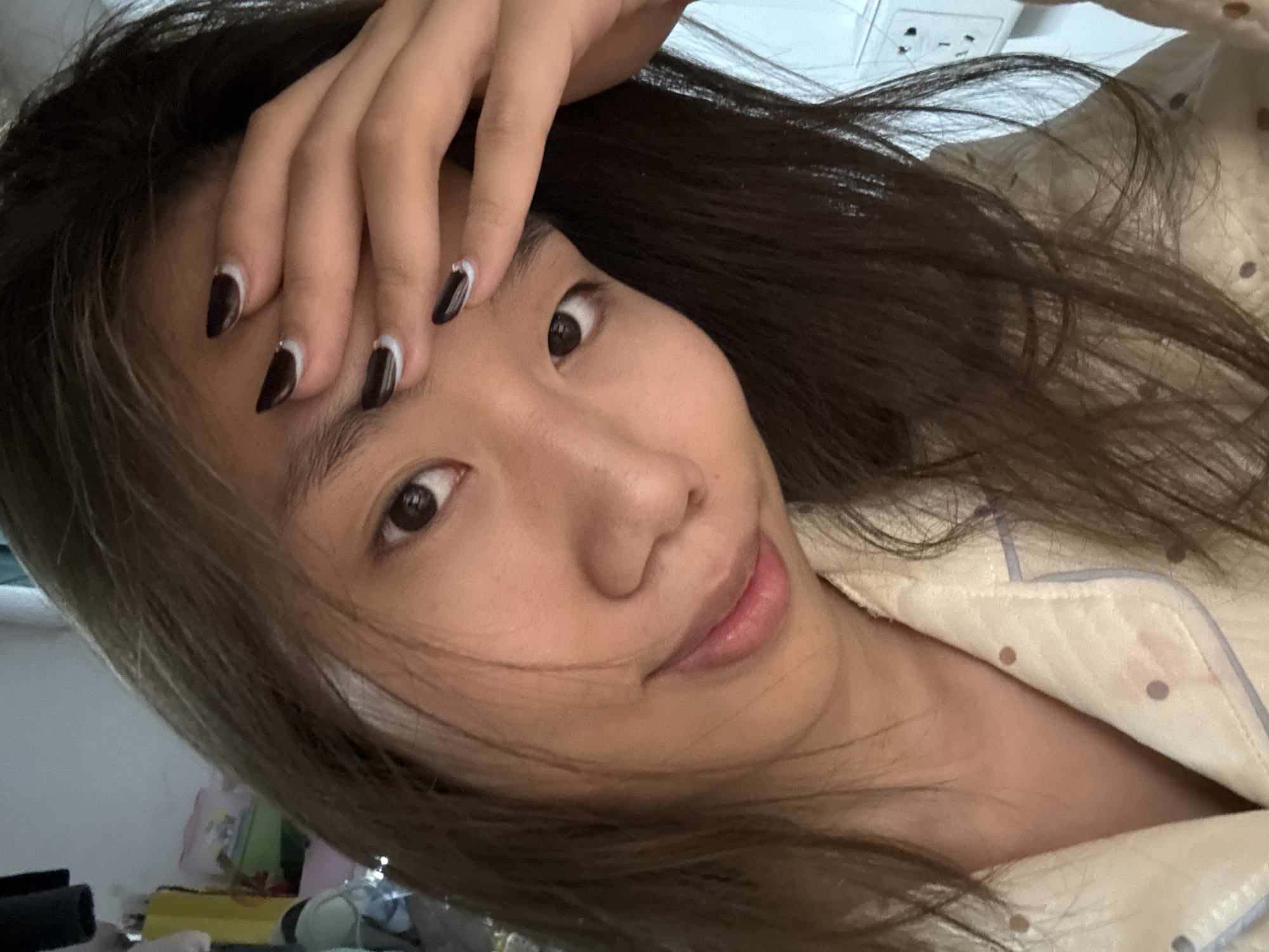 A selfie photograph shows a woman with long brown hair and brown eyes, wearing a pajama top. Her left hand is positioned on her forehead, and she has dark nail polish with a lighter accent at the base of her nails. The location is not discernible from the image alone. The Nan'an District, China location cannot be determined.