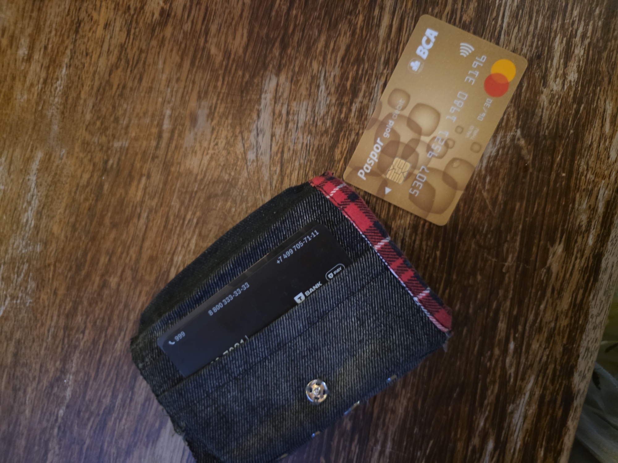 A black denim cardholder with red plaid trim is on a wooden surface. A dark-colored card is partially visible inside the cardholder. A gold-colored debit card with the logo "Paspor gold debit" and "BCA" is on the wooden surface next to the cardholder. The debit card has the number "5307 9521 1980 3196" and an expiration date of "06/30". The scene is likely indoors, given the lighting. The location is not specified beyond the general region of North Kuta, Indonesia. There are no people or actions depicted.