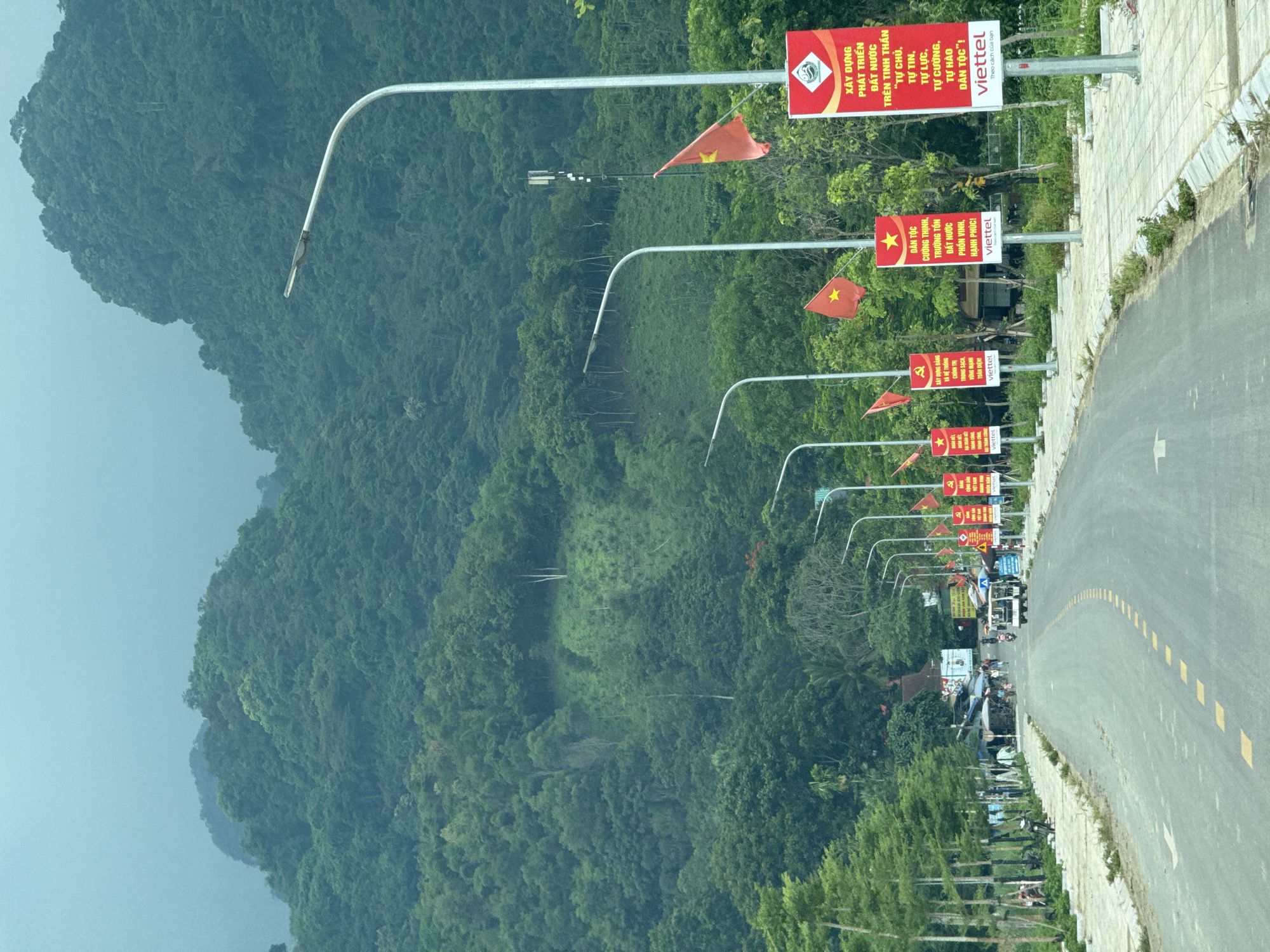 The image depicts a road leading through a verdant landscape in Cẩm Tú Commune, Vietnam. Lush green mountains dominate the background, shrouded in a light mist. Along the right side of the road, a series of lampposts are adorned with Vietnamese flags and red banners featuring Vietnamese text and imagery, including a star and the Viettel logo. A few people are visible on the side of the road near stalls and vehicles. A white electric vehicle is driving away from the camera. The road itself is asphalt with yellow dashed lines marking the lanes. Trees line the left side of the road, with some smaller trees and decorative plantings along a paved pedestrian walkway. The overall scene suggests a rural or semi-rural setting within a mountainous region, likely during overcast weather.