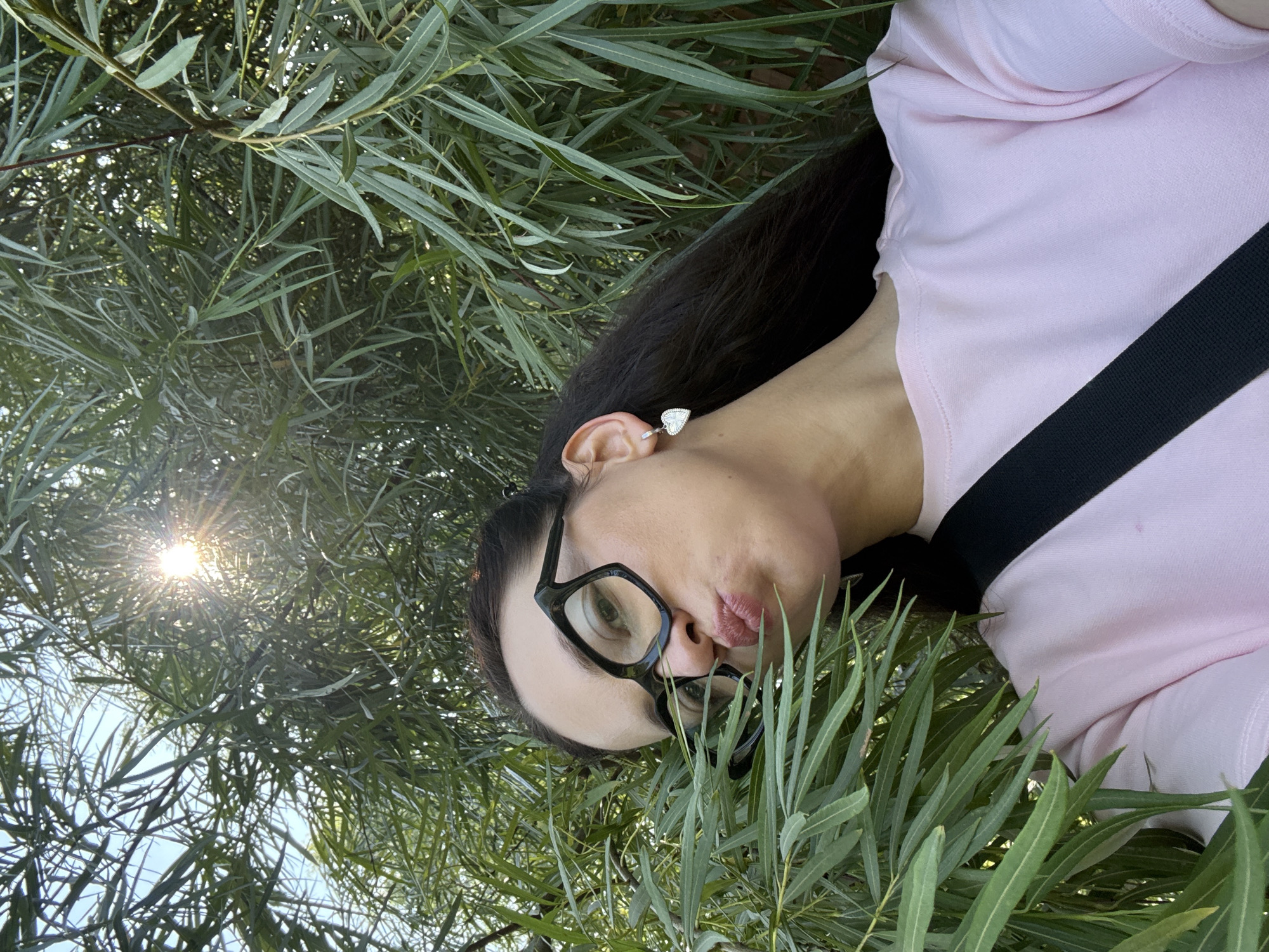 A woman is pictured from the chest up, looking out from behind dense green foliage. She is wearing large, dark-rimmed glasses and a pink crew-neck sweatshirt with a black strap across her chest. Her lips are pursed. The sun is visible through the leaves, casting rays of light. The image appears to be taken outdoors, with the foliage suggesting a natural setting. The location is identified as Chaikovsky, Russia.