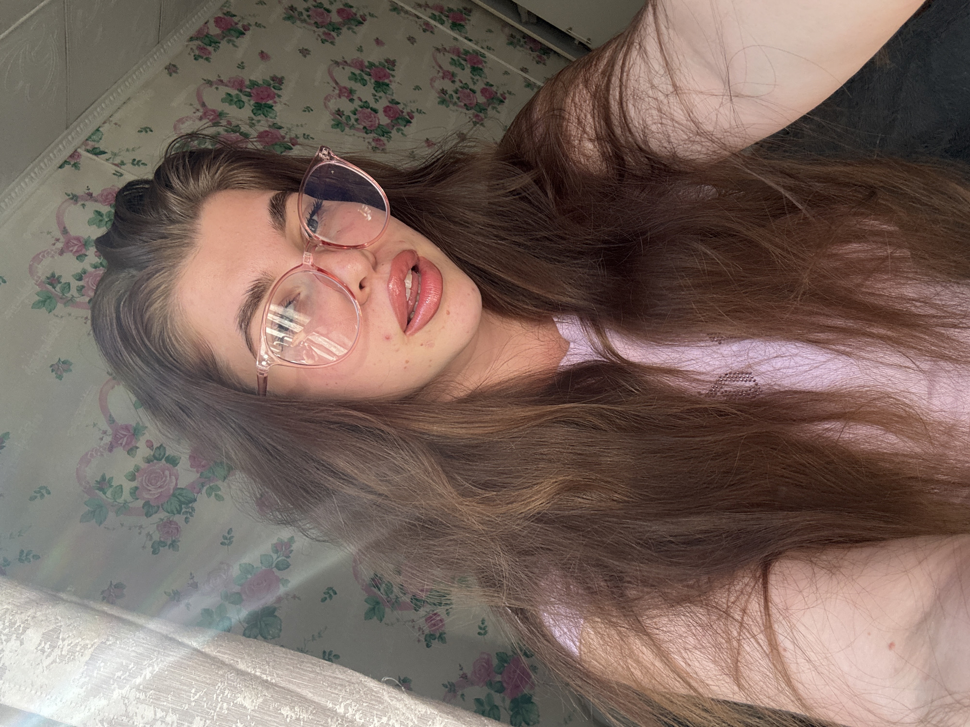 The image shows a close-up, high-angle, selfie-style shot of a young woman with long, brown hair. She is wearing pink-rimmed glasses and a light purple t-shirt. Her lips are slightly parted, and she has a subtle sheen on them. Her skin appears to have some blemishes and freckles. The background features a floral wallpaper with pink roses and green leaves, and a textured white curtain on the left side. Sunlight is streaming in from the left, creating lens flares and highlighting the textures in the image. The scene appears to be indoors, likely a bedroom or living area. There is no indication of the location being specifically in the city of Мещёры, Russia.