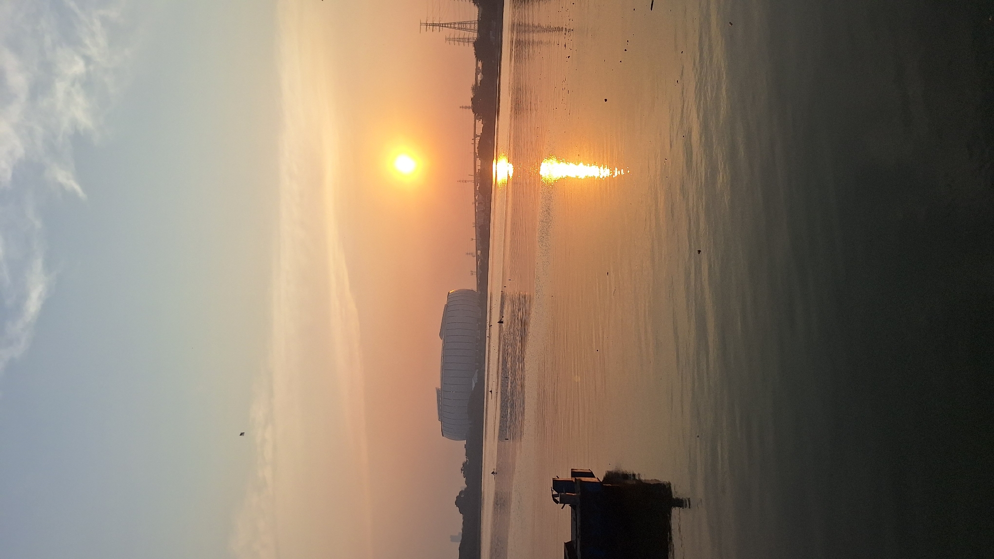 This is a vertical, eye-level, medium shot photograph taken at sunset over a body of water, likely in North Jakarta, Indonesia. The sky is a gradient of soft blue at the top transitioning to a warm orange and yellow near the horizon, with scattered wispy clouds. A bright, setting sun is visible in the upper right quadrant of the image, casting a strong, shimmering reflection on the water. In the distance, across the water, a large, modern, oval-shaped stadium with a ribbed exterior is prominent. To the right of the stadium, several tall, latticed electricity pylons are visible against the twilight sky. A small, dark boat is positioned in the lower left foreground, partially submerged and reflected in the water. Several indistinct figures are present in the water, some appearing to be in small boats or swimming. The water itself is calm, with gentle ripples reflecting the sunset colors. Small specks, possibly debris, are scattered across the water's surface.