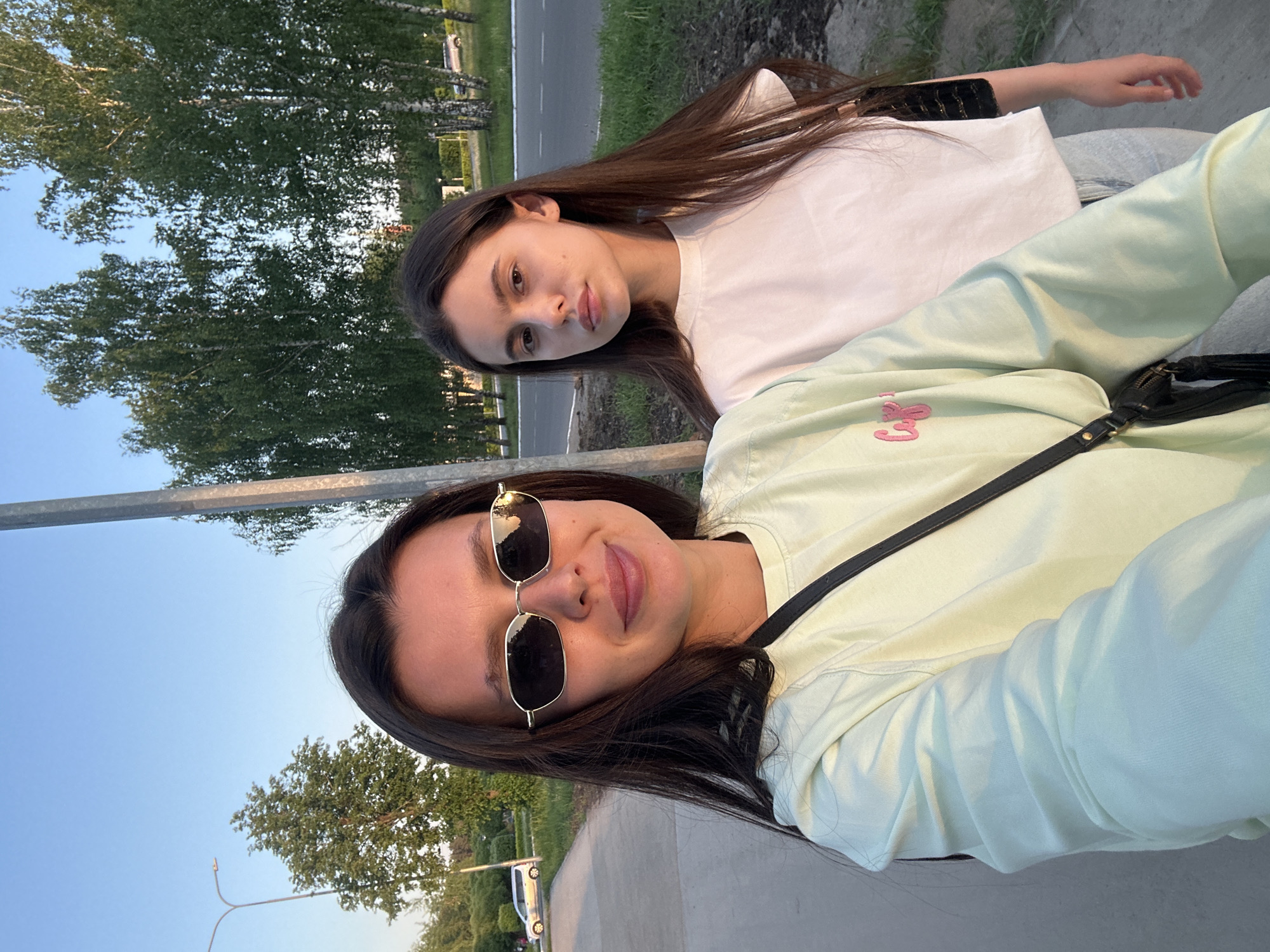 Two young women are positioned close together, likely posing for a selfie. The woman in the foreground wears sunglasses and a light green sweatshirt with a small pink design, possibly on the upper left chest. A black purse strap is visible across her body. The woman in the background is in a white t-shirt. The background reveals a street, trees, and blue sky, suggesting an outdoor setting in Chaikovsky, Russia.