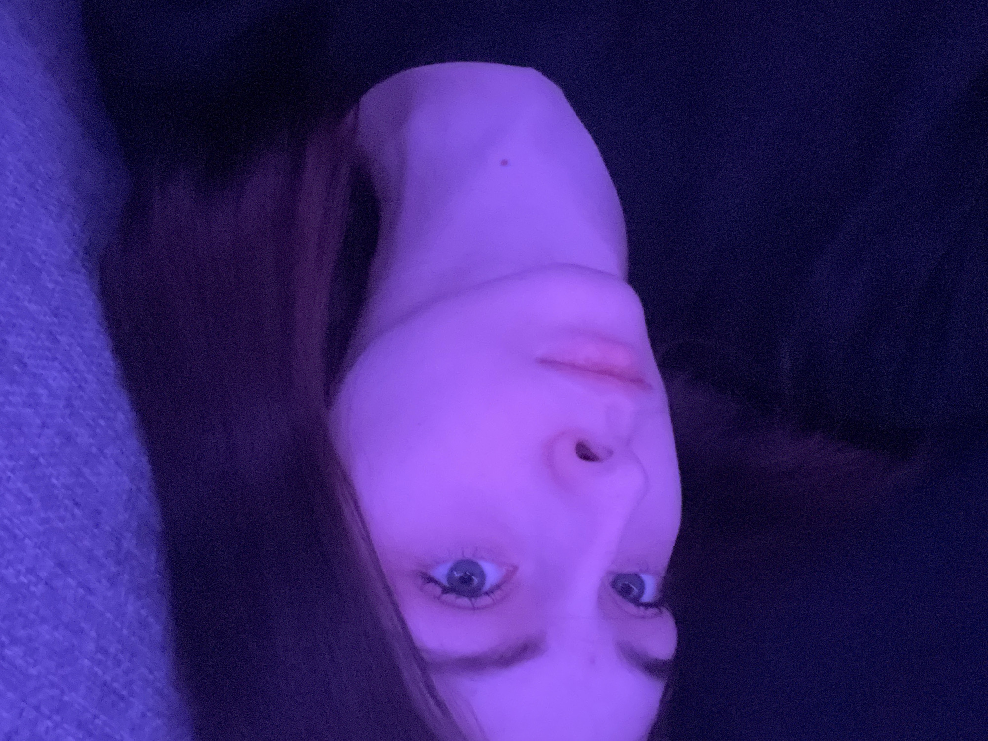 A close-up, dimly lit, rotated image shows a person's face with blue eyes. The lighting appears to be a purple hue, and a section of dark hair is visible on either side of the face. The background is dark and indistinct. No evidence suggests this photo was taken in Saint Petersburg, Russia.