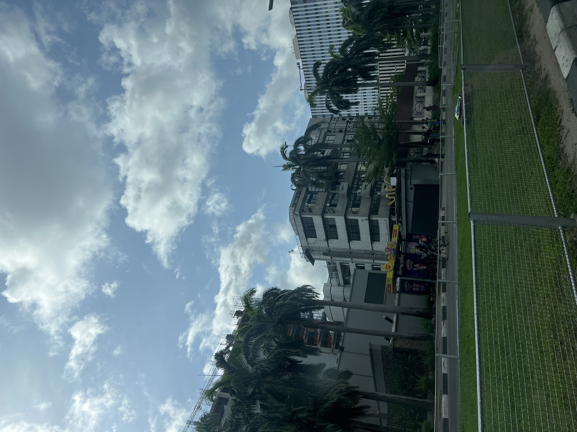 The image captures a cityscape in Ikoyi, Nigeria, under a sky dotted with fluffy white clouds. Several multi-story buildings are visible, some with palm trees in front of them. A metal fence runs alongside a grassy area, potentially marking a perimeter. Several motorcycles are parked near a roadside kiosk. Some people can be seen standing near the kiosk.