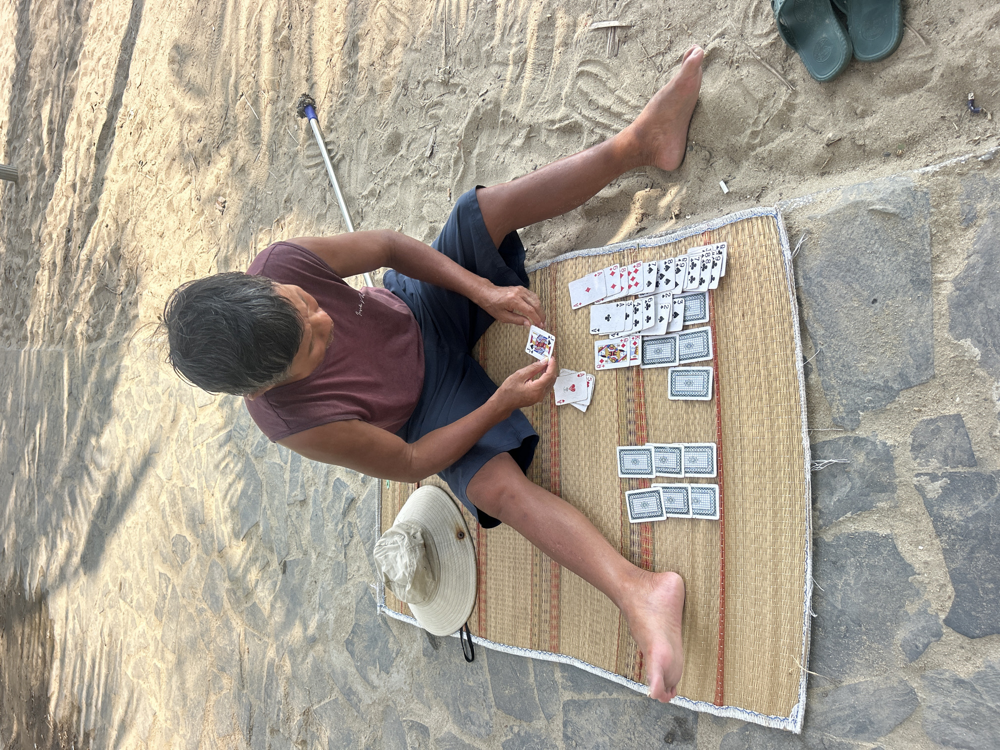 A man is playing solitaire on a woven mat on the beach at the city of Bắc Nha Trang, Vietnam. The mat is placed on a combination of sand and stone. The man is sitting cross-legged, wearing a maroon t-shirt and navy shorts. He is holding playing cards in his hands. The layout of the game is visible on the mat. A sun hat and a pair of sandals are positioned beside the mat. A long-handled tool lies in the sand.
