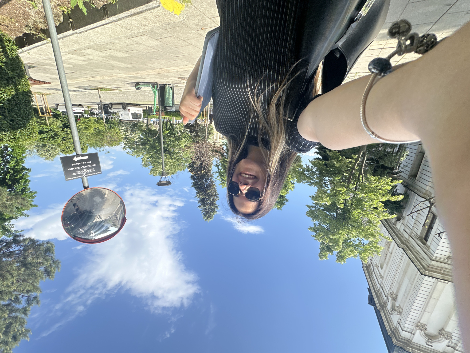 A selfie shows a woman with sunglasses and long brown hair standing outdoors on a sunny day. The background includes a clear blue sky with some clouds, trees, a sidewalk, a building, and a street sign.
The street sign has the words "Physic Garden" written on it. A trash bin is near the street sign. The reflection in a convex mirror shows details of the surroundings.
Based on the signage, the location is likely near or at the Physic Garden in Sofia, Bulgaria.