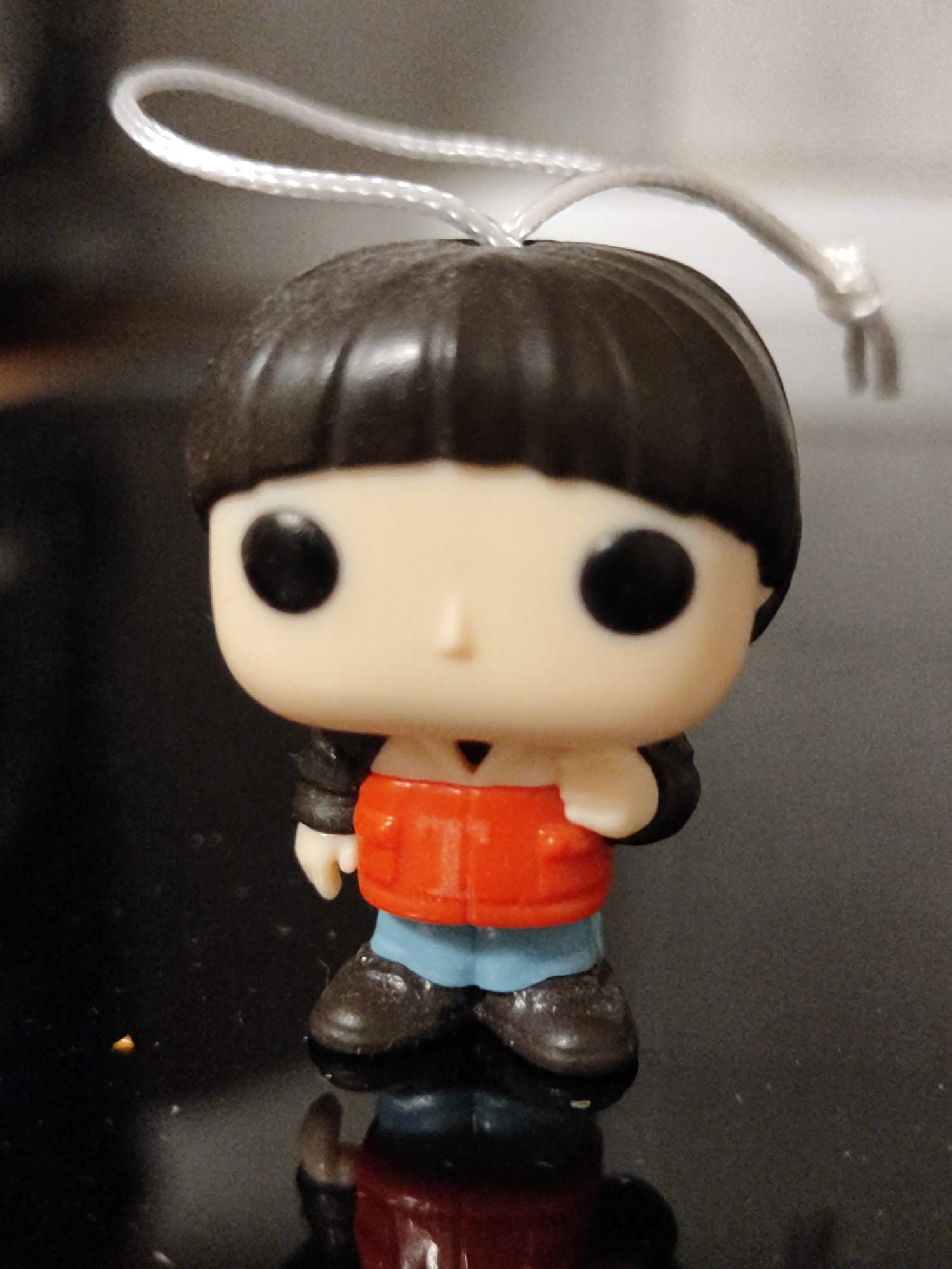A stylized figurine, typical of a Funko Pop design, is prominently featured in a close-up shot. The figure has a pale complexion, large black circular eyes, and a small, subtle nose. Its hair is dark brown, cut in a bob style with bangs. A silvery string loop is attached to the top of its head, suggesting it functions as a keychain or ornament.
The character is dressed in a vibrant red upper garment, possibly a jacket or vest, paired with light blue trousers and dark brown or black shoes. The figure stands on a highly reflective, dark surface, which creates a distinct, mirror-like reflection of its lower half and feet. The background is softly blurred and appears as a neutral, dark gray to black expanse, indicative of an indoor setting.
There is no discernible activity taking place, as the image captures a static object. No text is visible anywhere on the figurine or in the background. The lighting is soft and consistent, illuminating the figure and its reflection. The image depicts a collectible toy. The location context for this media file is Vozdovac Urban Municipality, Serbia.