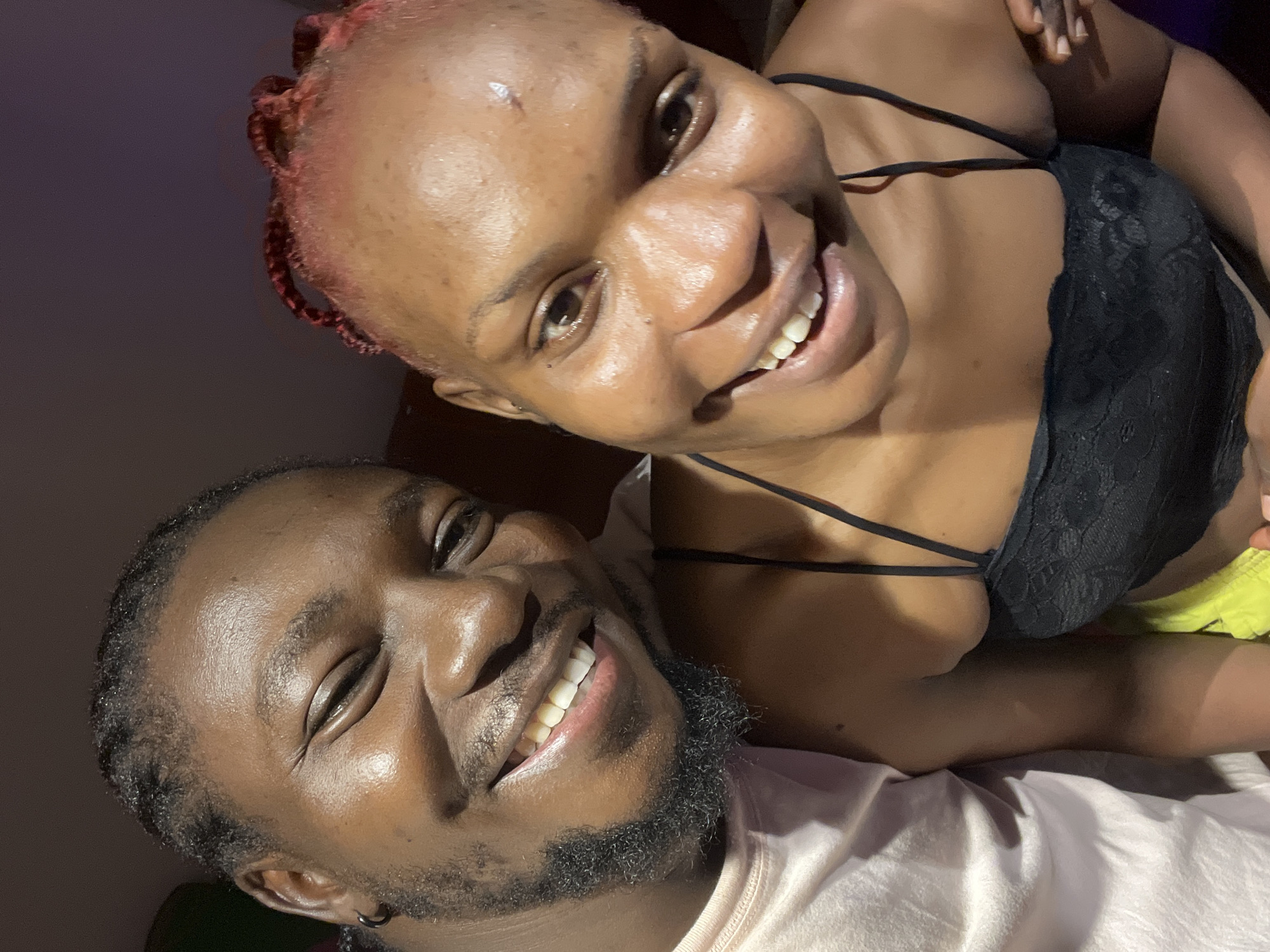 The image is a close-up selfie featuring a man and a woman, likely taken indoors. The man, on the left, has dark skin, braided hair, a beard, and is wearing a light-colored t-shirt. He is smiling broadly, with his eyes partially closed. The woman, on the right, also has dark skin, with her hair styled in braids with reddish highlights. She is wearing a black lace camisole and is also smiling. Their faces are very close together, suggesting intimacy. The background is dark and out of focus, with a hint of a purple hue. The lighting appears to be artificial. There are no discernible landmarks or clues in the image that would place the location specifically in Jos, Nigeria.