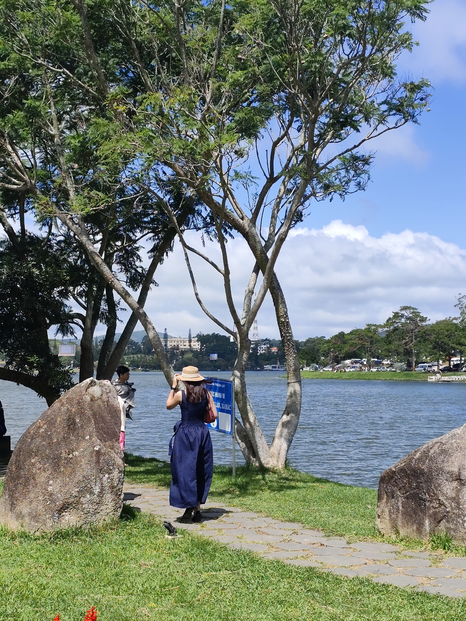 The image captures a sunny day scene at Xuan Huong Lake in Da Lat, Vietnam. In the foreground, a person wearing a dark blue dress and a straw hat stands on a stone-paved path, facing away from the camera and towards the lake. They are holding a red bag in their right hand and adjusting their hat with their left. To the left of the path, a large, rough-textured rock sits on a patch of green grass. Behind the person, a body of water, presumably Xuan Huong Lake, stretches out, with its surface rippling gently.
In the middle ground, on the opposite bank of the lake, a line of trees forms a green border. Beyond the trees, a large, light-colored building with a distinctive architectural style, possibly a historical landmark or hotel, is visible against a bright blue sky dotted with white clouds. The background also shows more trees and some distant structures, suggesting the presence of the city surrounding the lake.
To the left of the main subject, partially obscured by trees, is another person, possibly a child, wearing a light-colored top and dark bottoms, looking towards the lake. A blue signpost stands near the main subject, with text written in Vietnamese, indicating information or regulations for the area. The overall atmosphere is peaceful and scenic, typical of a popular tourist destination like Xuan Huong Lake.