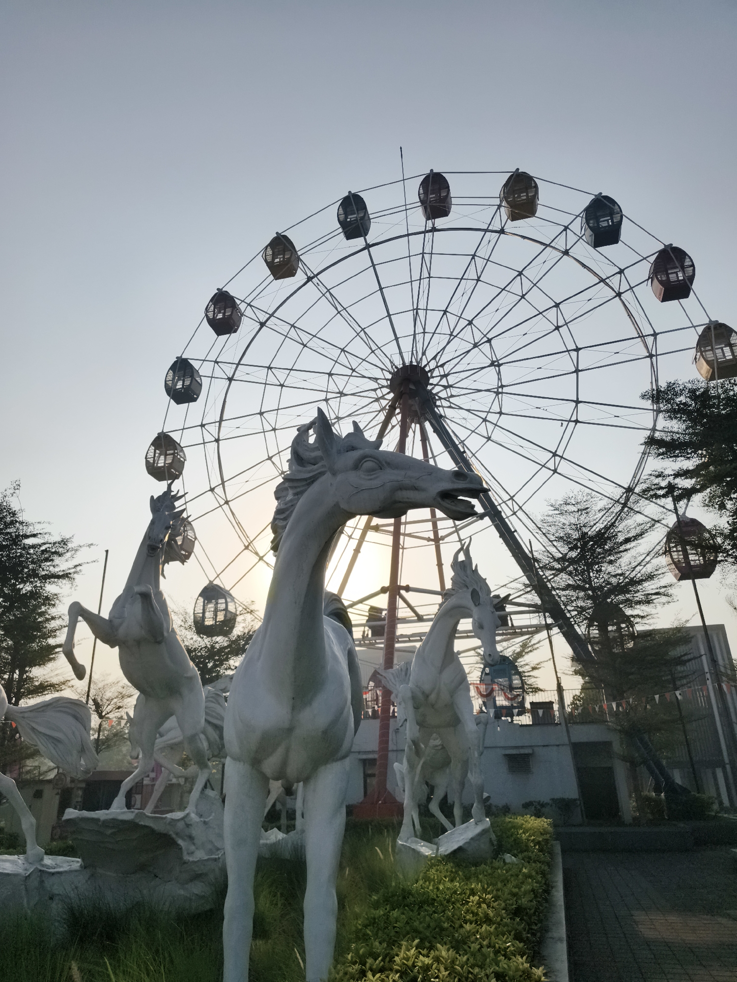 In Petiken, Indonesia, at what appears to be an amusement park or recreational area, a Ferris wheel stands prominently against a pale sky during twilight. The Ferris wheel is circular, with numerous passenger gondolas suspended around its circumference. In the foreground, several white horse sculptures are positioned, adding a decorative element to the scene. One sculpture in the immediate foreground depicts a horse's head and neck. Behind it, another horse sculpture is rearing up on its hind legs. Further back, more horse sculptures are visible. The surrounding environment includes trees, some of which are bare, and a building structure to the right. A paved pathway runs along the right side of the image. The lighting suggests it is either early morning or late afternoon.
