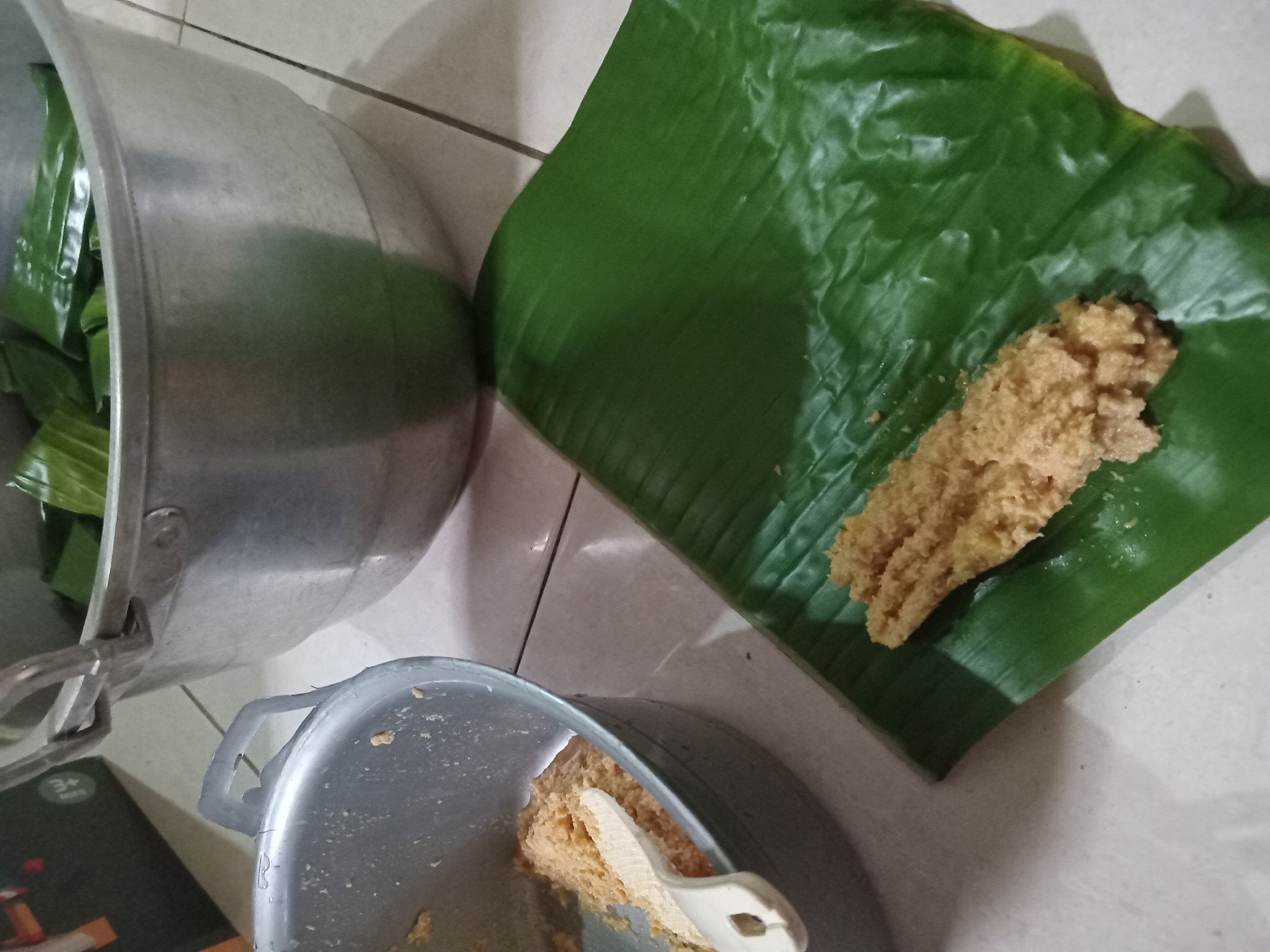 A close-up, high-angle shot shows a culinary preparation scene, likely in Claket, Indonesia. In the foreground, a large, vibrant green banana leaf is laid out on a tiled surface. On top of the leaf sits a portion of a light brown, grainy mixture, resembling a filling or batter. To the left, a metal pot contains a similar light brown mixture, with a white plastic spatula resting inside. In the background, a larger aluminum pot is filled with what appear to be more banana leaf-wrapped items, suggesting a larger batch of food preparation. The overall impression is one of domestic food production, possibly for a traditional Indonesian dish.