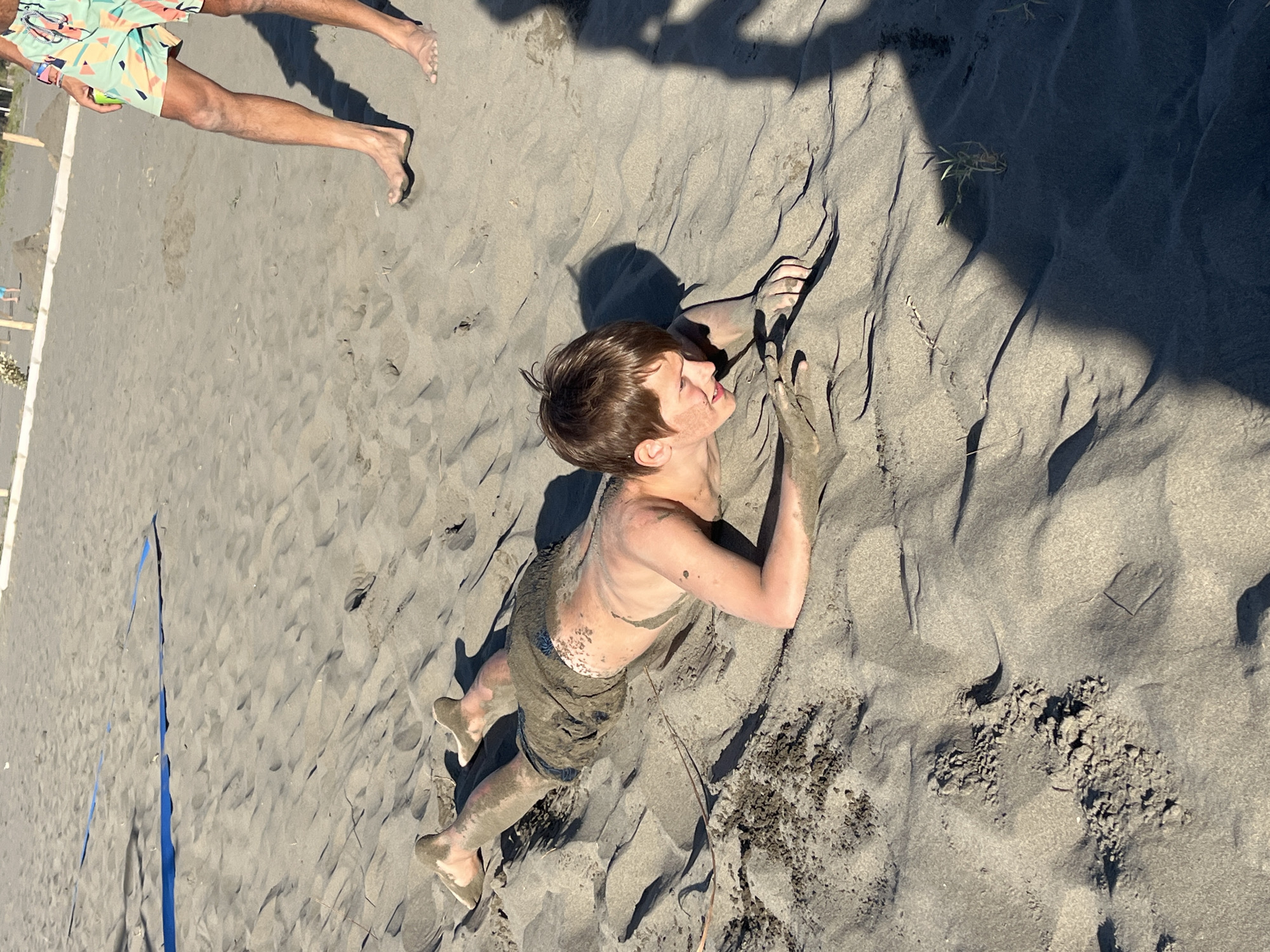 A young Caucasian male, appearing to be in his early to mid-teens, is lying prone on a dark sand volleyball court. He is covered in sand from his torso to his feet. He is wearing dark-colored shorts. His right arm is extended forward, and his left arm is bent, with his hands resting in the sand. His facial expression is difficult to discern clearly. In the background, another person's legs are visible, wearing colorful patterned shorts. The sand surface shows ripples and indentations, suggesting recent activity. The court has blue boundary lines visible in the upper left portion of the image. The overall environment suggests an outdoor recreational area, likely a beach or sports facility.