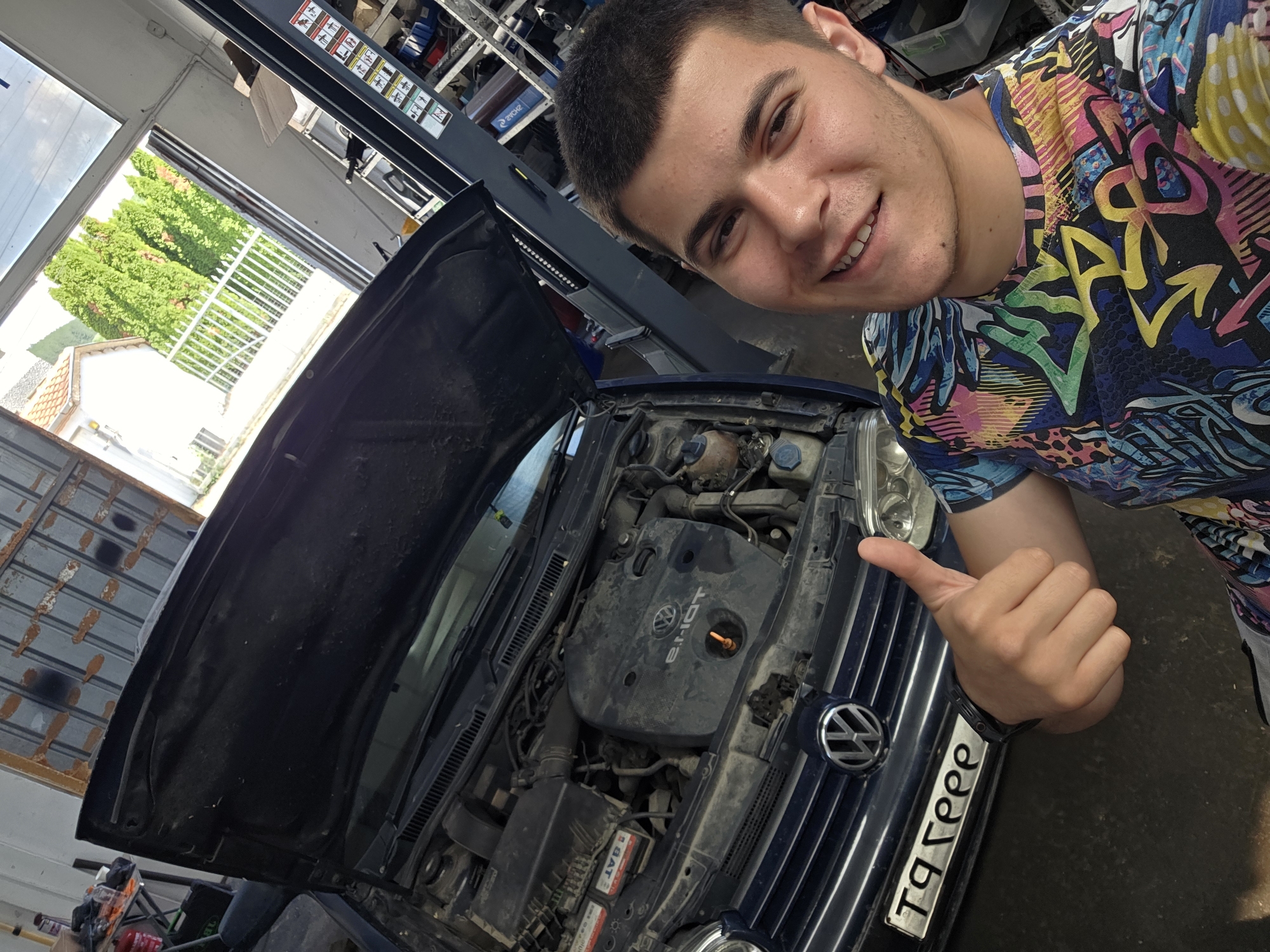 A young man with short brown hair, smiling and giving a thumbs-up gesture, is taking a selfie in front of a blue Volkswagen with its hood open. The car appears to be in a garage or workshop, indicated by the presence of a car lift in the background, shelves with equipment, and the overall industrial setting. The man is wearing a colorful t-shirt with graffiti-style patterns and athletic shorts. The license plate of the car is visible and reads "T9 GEEE". The environment suggests a repair shop, possibly located in or near Dragichevo, Bulgaria, as indicated by the context. The scene captures a moment of satisfaction or completion, possibly after a car repair.