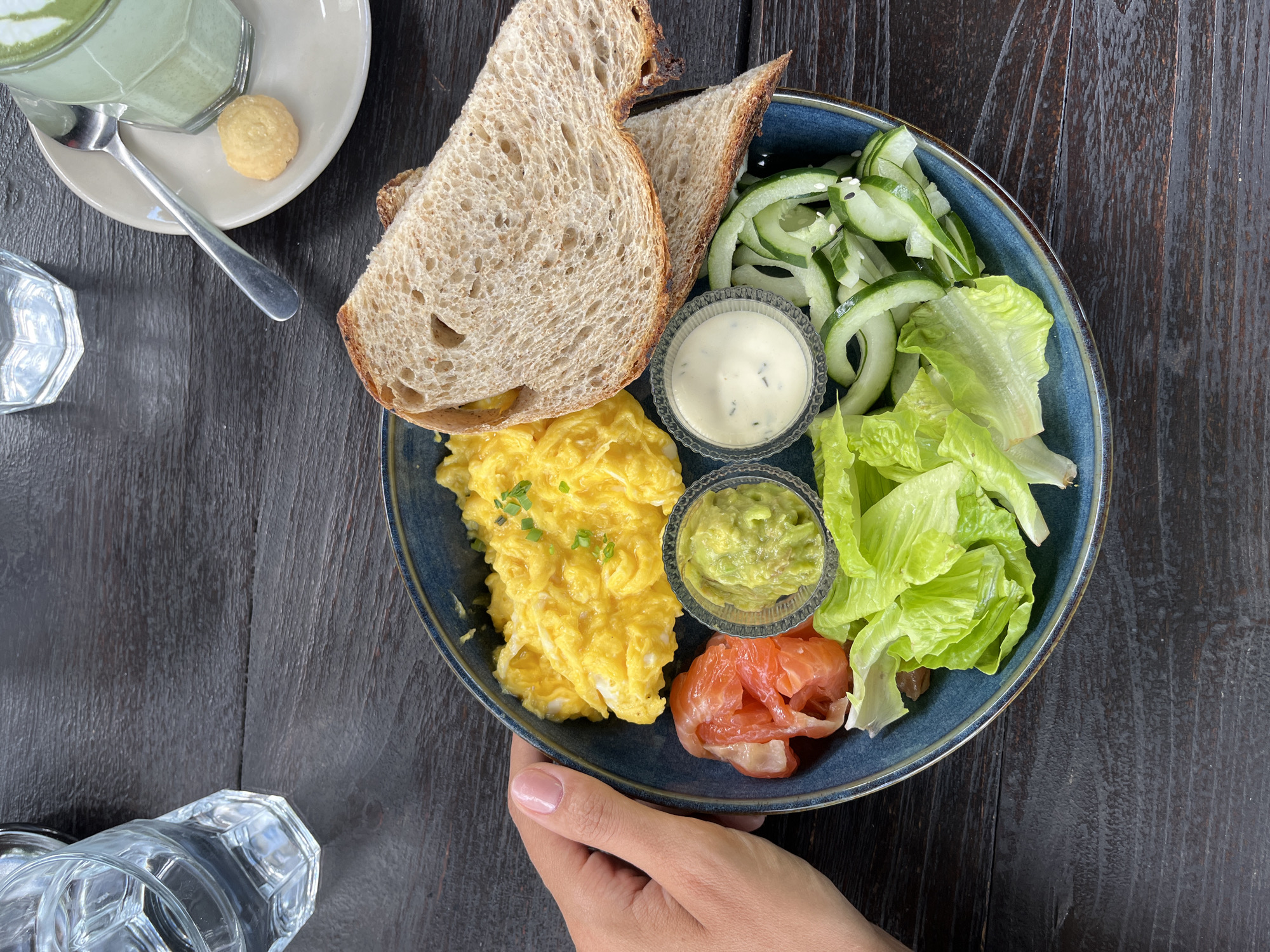 The image depicts a breakfast scene, likely in Kuta Selatan, Indonesia, given the geotags. The scene is shot from an overhead perspective on a dark wooden table. A person holds a blue ceramic plate with a breakfast composition. On the plate, there are scrambled eggs, slices of toasted bread, rolled cucumber slices with sesame seeds, lettuce leaves, a small container of guacamole, a small container of cream sauce, and a rolled piece of salmon. To the left, there is a glass of matcha latte with a small cookie on the saucer. At the bottom left corner, a drinking glass is visible. The overall composition suggests a bright, daylight setting.