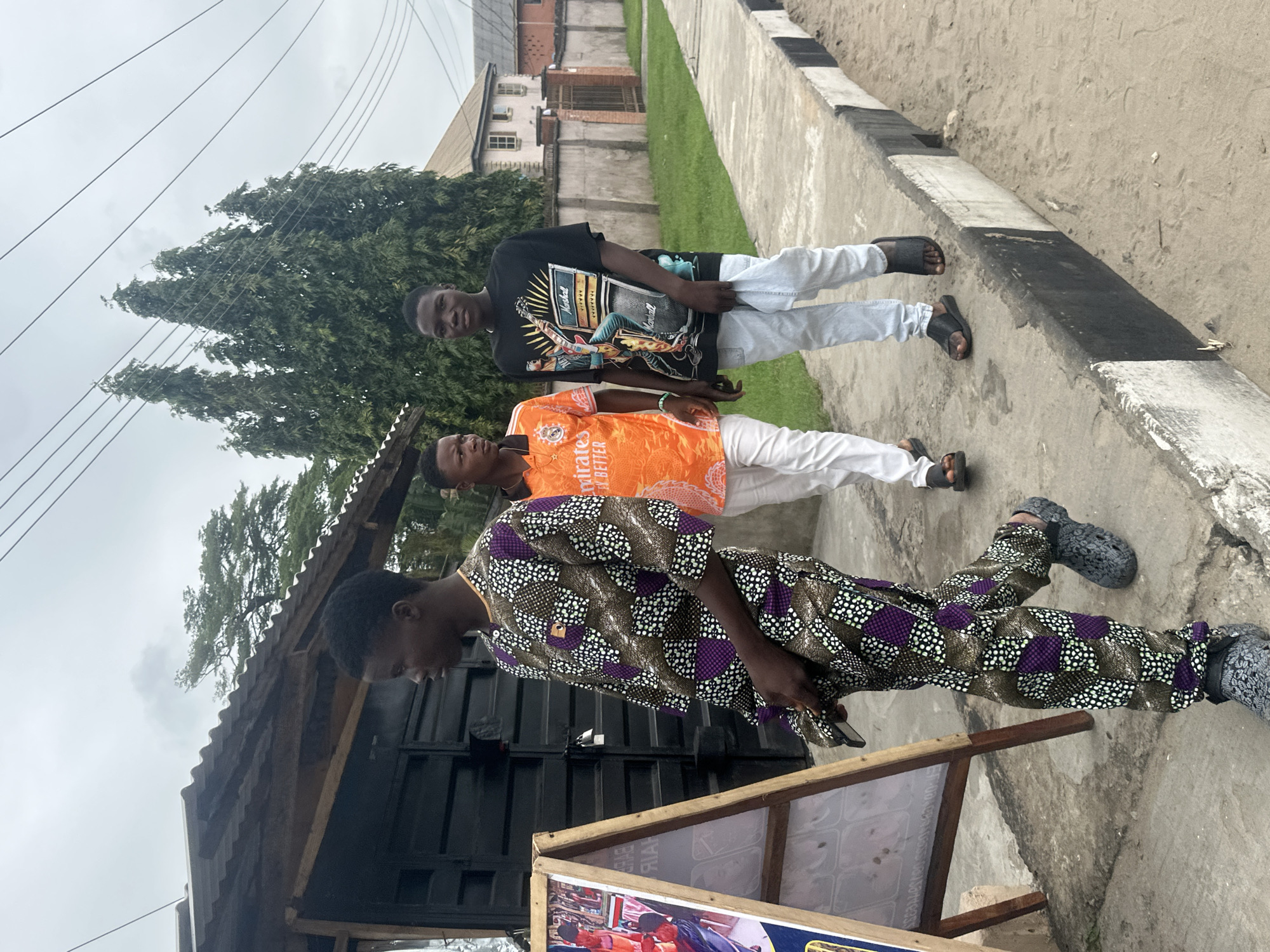 Three young Black males are walking on a concrete sidewalk next to a sandy dirt road. The male in the foreground, on the left, is wearing a traditional Nigerian outfit consisting of a patterned brown and purple tunic and matching pants, along with gray Crocs. He is looking to his left and holding a phone in his right hand. To his right, another male is walking and wearing an orange t-shirt with white writing and white pants, also wearing black sandals. Behind them, the third male is walking and wearing a black t-shirt with a graphic print and light blue jeans, with black sandals. He is holding a crumpled piece of white fabric in his left hand.
To the left of the sidewalk, there is a black corrugated metal structure with a corrugated metal roof, and a wooden easel with a poster on it. The poster is partially obscured and shows images of people, likely for a business such as a salon or barber shop. Behind the metal structure, there are tall green trees. Further down the street, there are houses and fences. The sky is overcast and gray, indicating a cloudy day. The scene appears to be in a residential or commercial area of Badagry, Nigeria, given the architecture and vegetation.