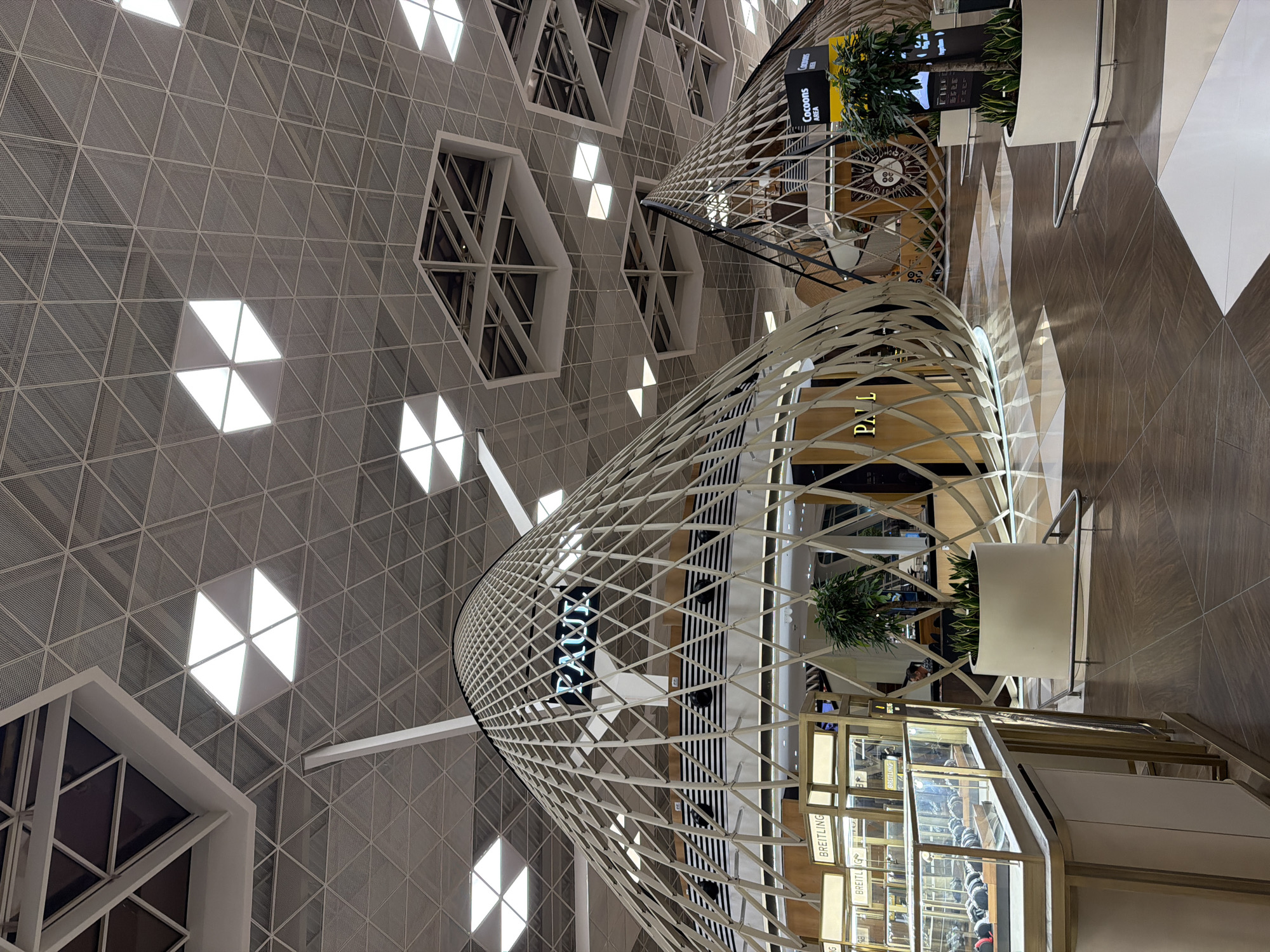 The image shows the interior of an airport terminal, likely Heydar Aliyev International Airport in Khazar Raion, Azerbaijan. The architecture is modern and features a distinctive geodesic dome-like structure with a latticed facade housing shops. One prominent sign reads "PAUL". In the foreground, a display case filled with watches is visible, with a sign indicating "BREITLING". Other shops, plants in large pots, and seating areas are also present. The ceiling is a complex structure of intersecting triangular panels and recessed lighting. The floor is tiled with a geometric pattern in shades of brown and white. The overall impression is one of a spacious and contemporary airport environment.