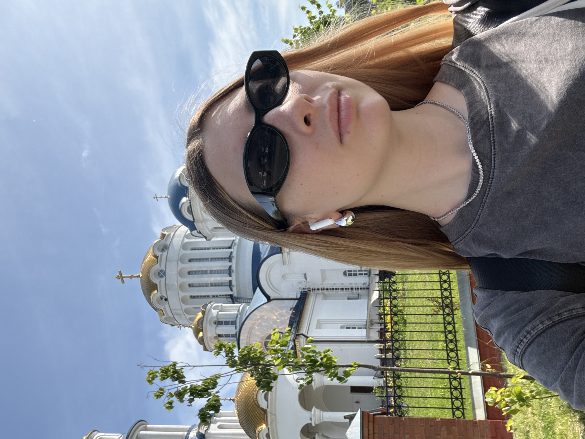 This is a selfie of a young woman with long blonde hair, wearing black oval sunglasses, silver hoop earrings, and a grey t-shirt. She is standing outdoors in front of a large, white Orthodox church with golden domes and blue accents. A small tree with green leaves is in the foreground, and a black metal fence separates the church grounds from a grassy area. The sky is blue with some clouds. The church appears to be the Church of the Holy Tsar Martyrs and Confessors in Moscow, Russia.