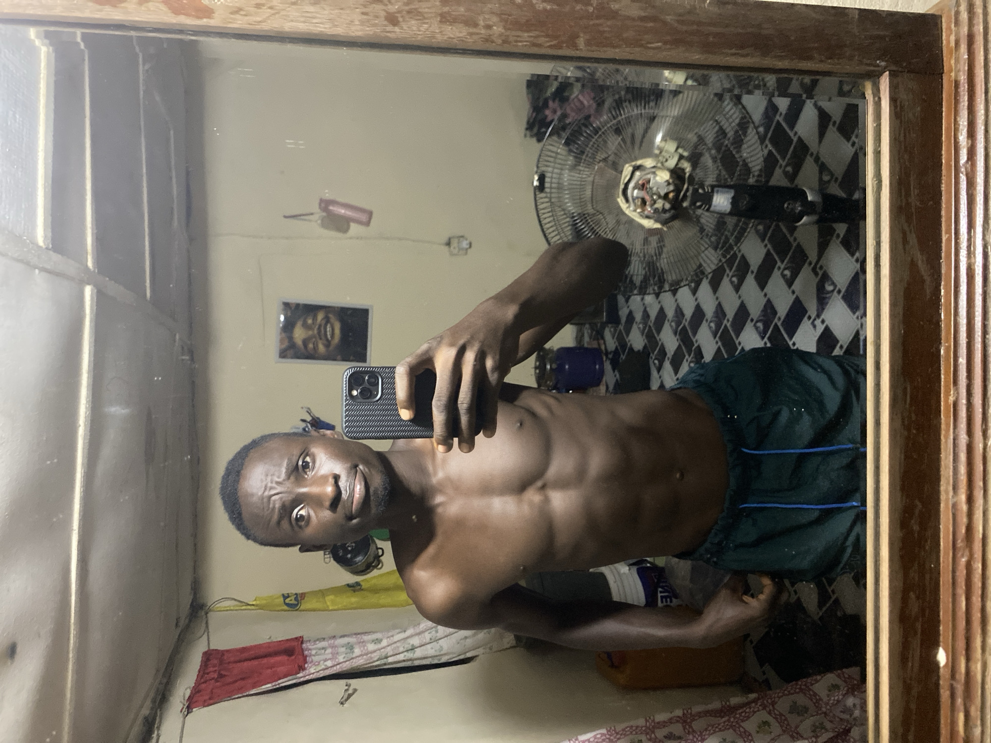 A young Black male, shirtless and wearing dark green shorts with blue drawstrings, stands in front of a mirror, taking a selfie with a smartphone. He has short, dark hair and a prominent brow. His torso is muscular, displaying visible abdominal muscles. The background, reflected in the mirror, depicts a sparsely furnished room. To the left, a red and white patterned cloth hangs on the wall, next to a yellow object and a large orange container. A framed picture of a smiling Black person is visible on the wall behind him. In the right side of the reflection, a black oscillating fan with a damaged grill stands on a checkered floor. The room appears to be in a state of disarray, with various objects and a general lack of tidiness. The lighting is dim, casting shadows and highlighting the textures of the room. The location is described as Barden Yana, Nigeria.