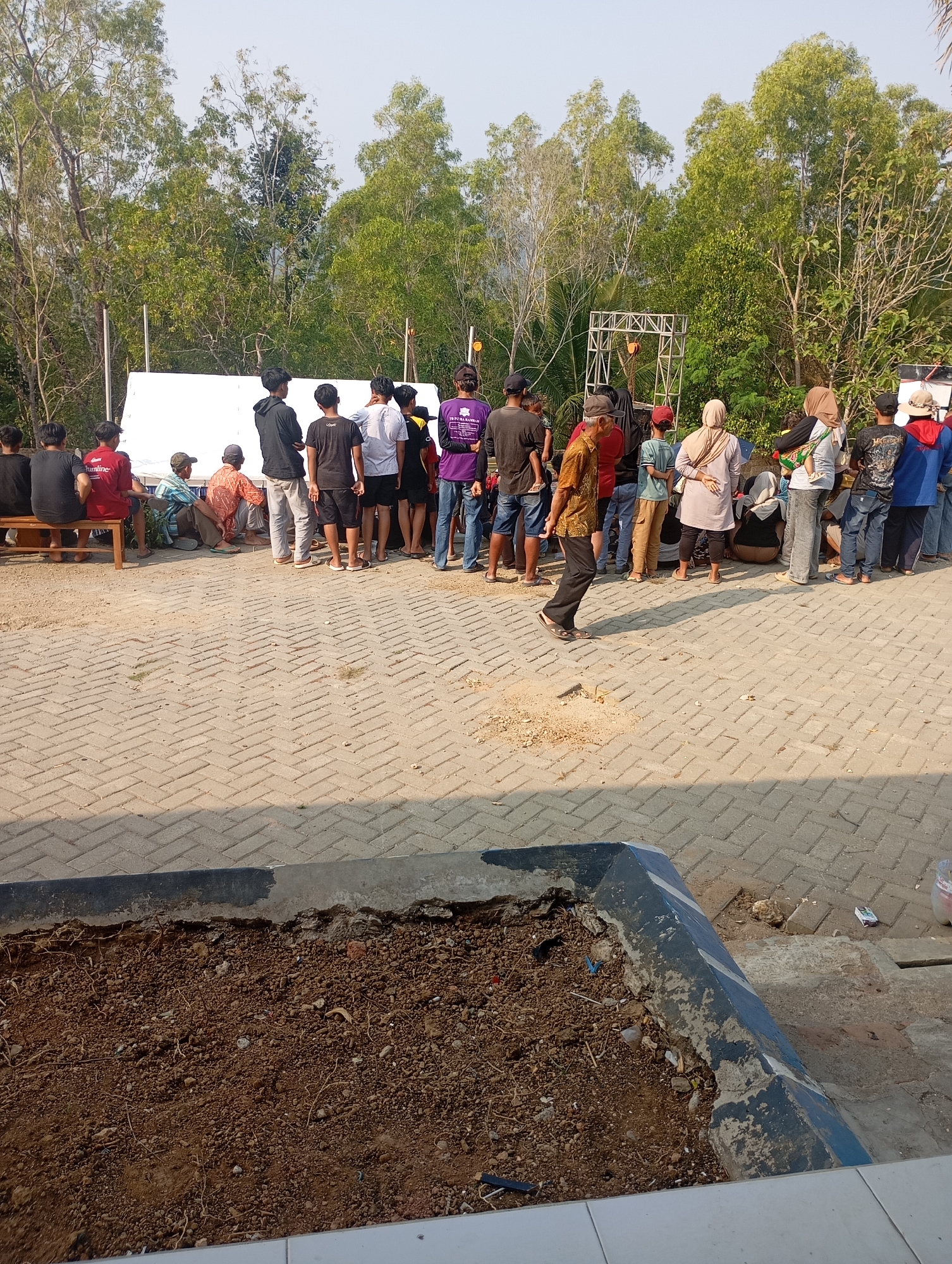 The image shows an outdoor gathering of approximately twenty-five people standing and sitting on a paved area in front of a white screen and a metal stage structure. The background consists of trees and a clear sky. In the foreground, there is a raised garden bed filled with soil and debris. The people are dressed in casual clothing, and some are looking towards the screen or stage. One person in the midground is walking away from the camera. The scene appears to be set in Ketepung, Indonesia, as indicated by the context.