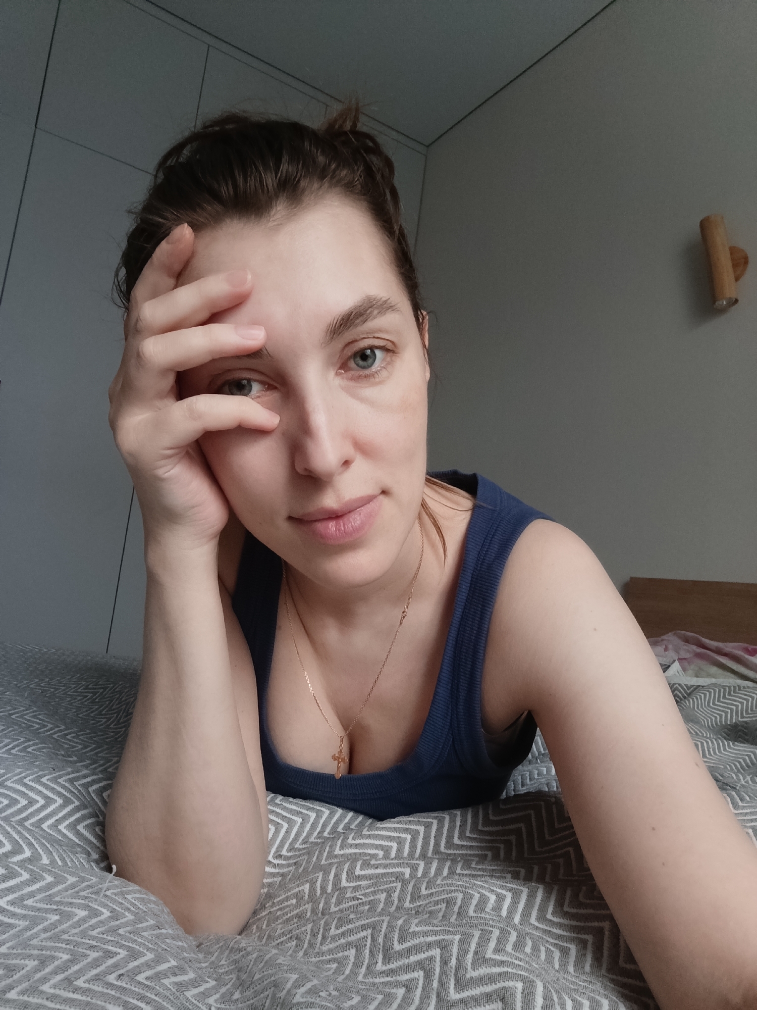 A young woman is lying on a bed with a gray and white chevron-patterned bedspread. She is wearing a blue tank top and a gold necklace with a cross pendant. Her dark hair is pulled back in a messy bun. She is resting her head on her hand, looking directly at the camera. The background consists of a light gray wall with a white cabinet on the left and a wooden light fixture on the right. There is no evidence to indicate the location of the image.