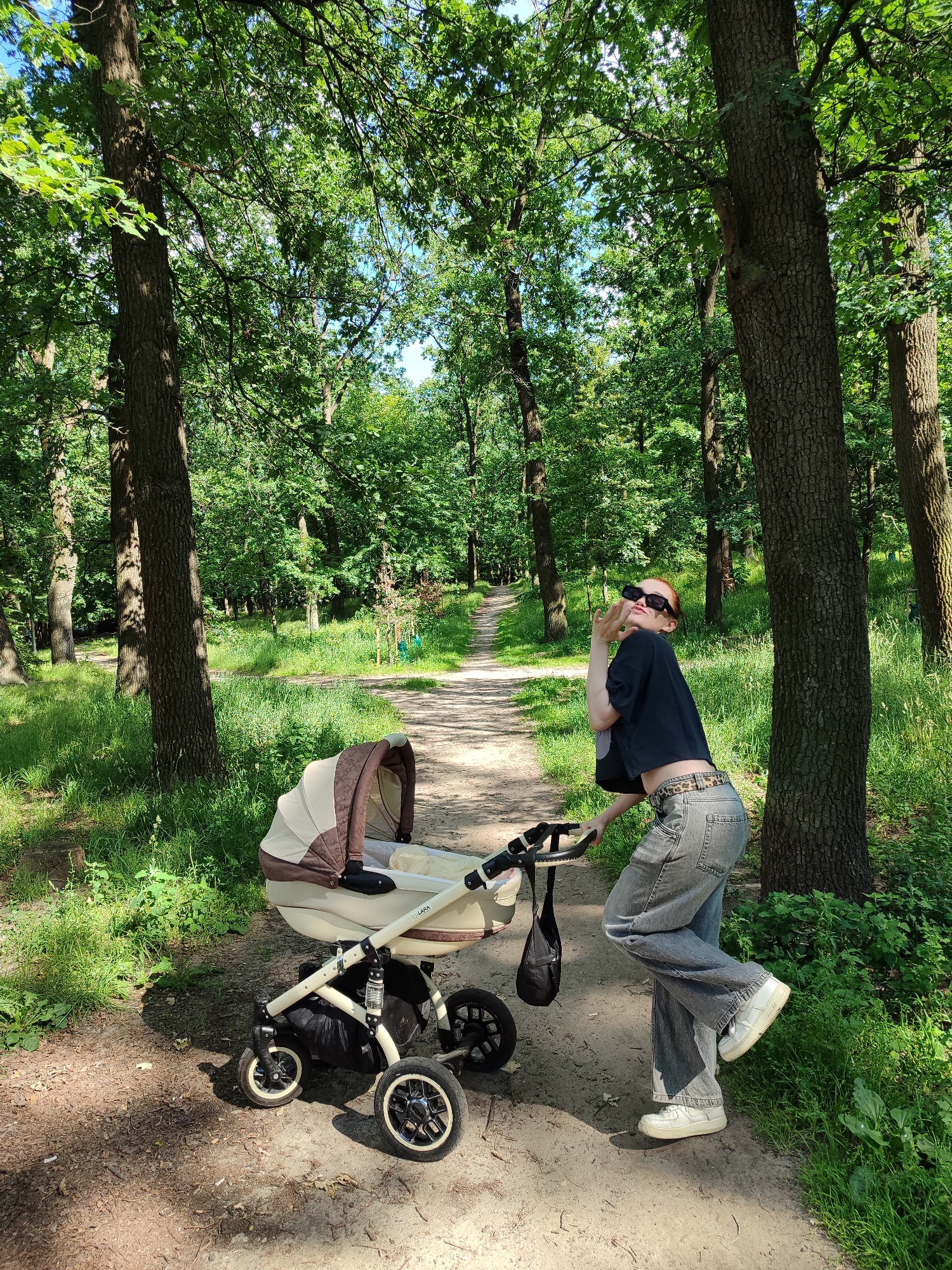In Kyiv, Ukraine, a young woman with red hair, wearing sunglasses, a dark cropped t-shirt, and grey jeans, poses playfully next to a beige and brown baby stroller on a dirt path in a park. The path winds through a dense forest with tall trees and lush green undergrowth. Sunlight filters through the canopy, creating dappled patterns on the ground. The woman is leaning on the stroller, blowing a kiss to the camera, while her other hand is raised in a peace sign. The stroller is modern and appears to be in good condition. The overall atmosphere of the image is cheerful and relaxed, suggesting a pleasant day out in nature.