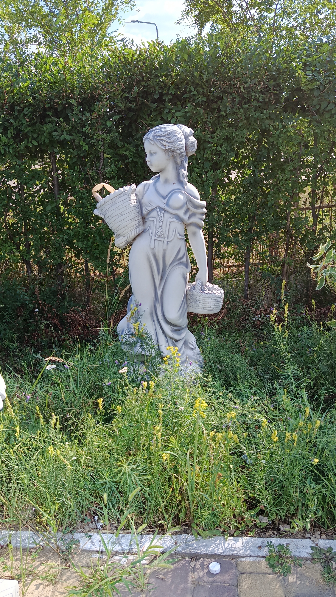 This is a full, eye-level, outdoor shot of a grey, plaster statue of a young woman holding two woven baskets. The statue is positioned in a garden, surrounded by lush green foliage and tall grass interspersed with small yellow and white wildflowers. Behind the statue, a dense hedge forms a wall of green, and in the upper portion of the image, a streetlamp can be seen protruding from behind the hedge. The foreground features a paved area with stone tiles, and scattered debris including a white plastic cap. The lighting suggests it is daytime with sunlight casting shadows. The scene appears to be in a public space or a well-maintained private garden within the city of Kostanay, Kazakhstan.