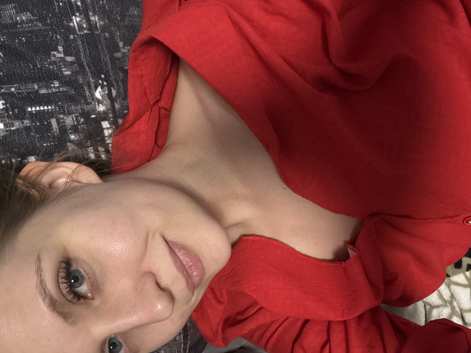 A woman with blue eyes and light brown hair is wearing a red hooded shirt and smiling slightly. She is lying on a patterned surface, possibly a blanket or sheet, that features a black and white cityscape design. The camera angle is slightly tilted. The location is unknown.