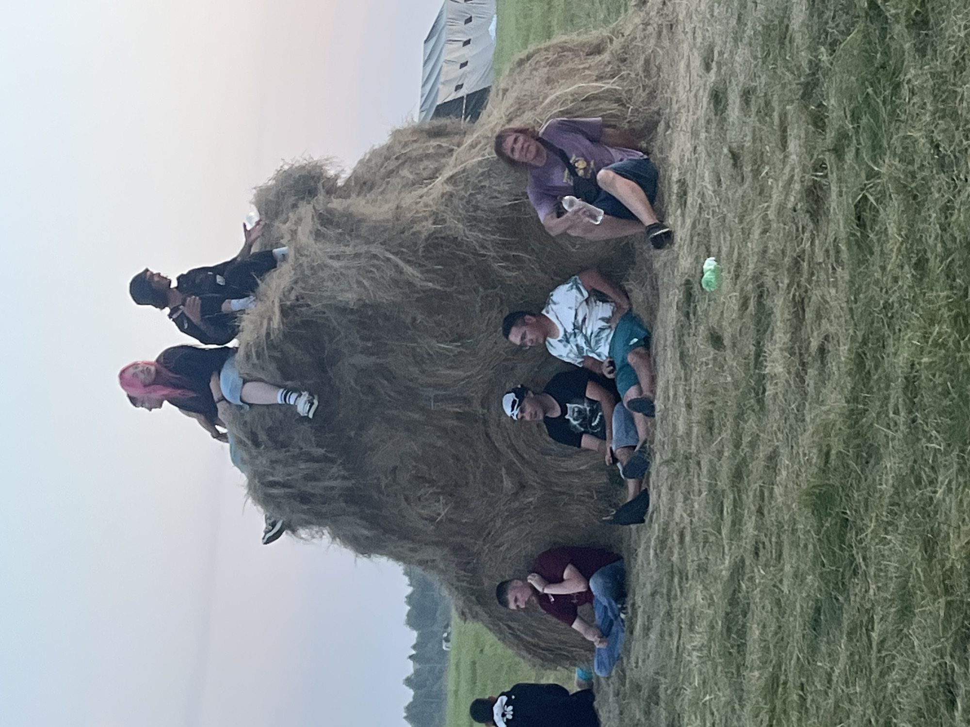 In Surakovo, Russia, a group of people are gathered around a large haystack. On top of the haystack, two individuals are seated. One has pink hair and is wearing a dark jacket and denim shorts. The other, to their right, is wearing a dark jacket and dark pants. Below them, seated at the base of the haystack, are five more individuals. On the left, a person in a maroon shirt and jeans sits cross-legged, holding their chin. Next to them, a person in a black t-shirt with a white graphic and dark shorts is seated. To their right, a person in a white and blue patterned t-shirt and turquoise shorts is also seated. Further to the right, a person in a purple t-shirt and dark shorts is holding a water bottle. On the far left, partially visible, is a person in a black jacket with a white design on the back. The environment is a grassy field with trees visible in the distance. A large tent structure is also present in the background.