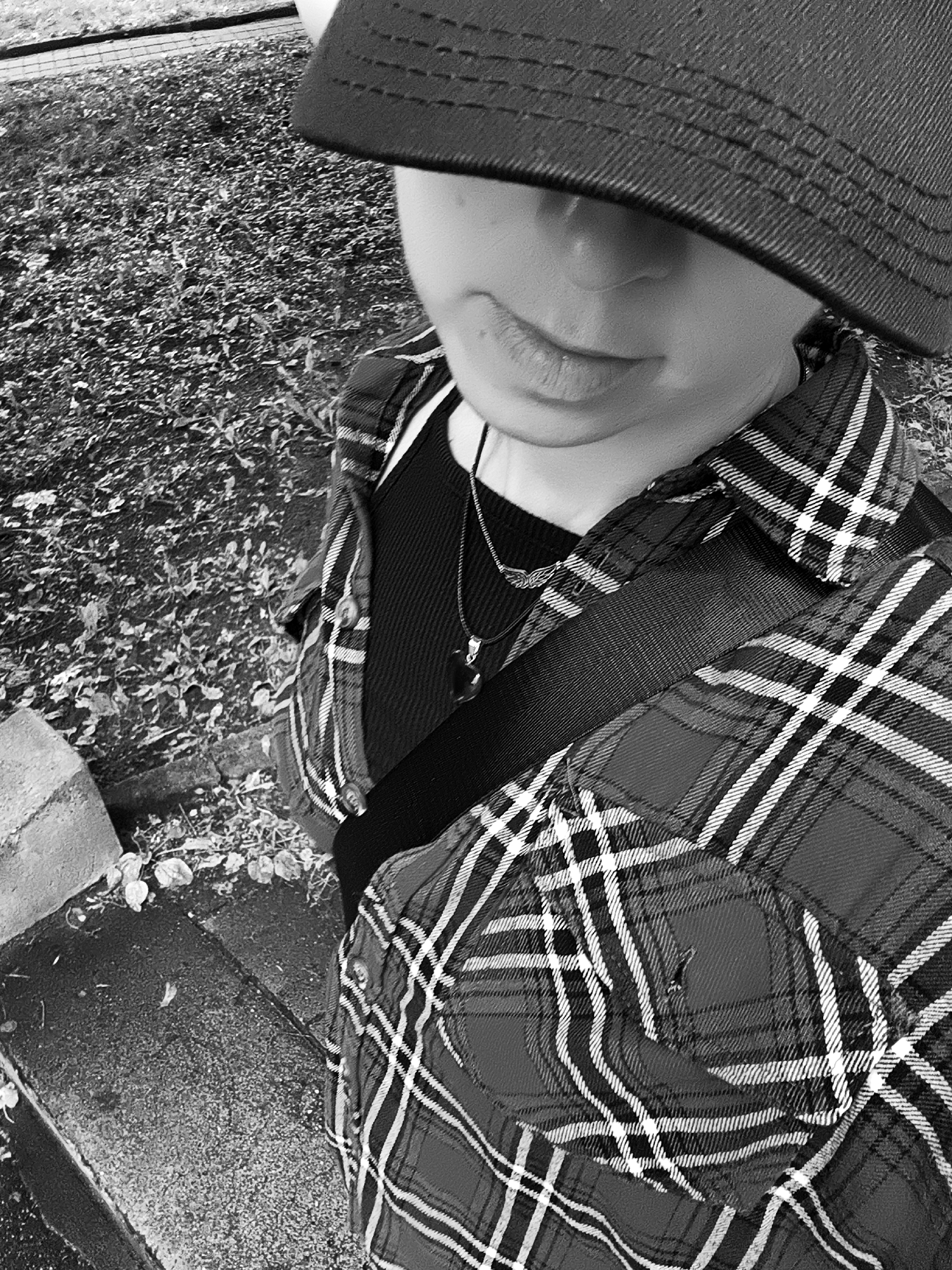 A black and white, high-angle, medium shot shows a person wearing a dark baseball cap, a black ribbed shirt, a red and black plaid shirt, and a necklace with a dark pendant. The person is standing outdoors in an area with patchy vegetation and a concrete structure. The composition focuses on the upper body and face of the subject, with the background blurred. The overall impression is informal and candid.