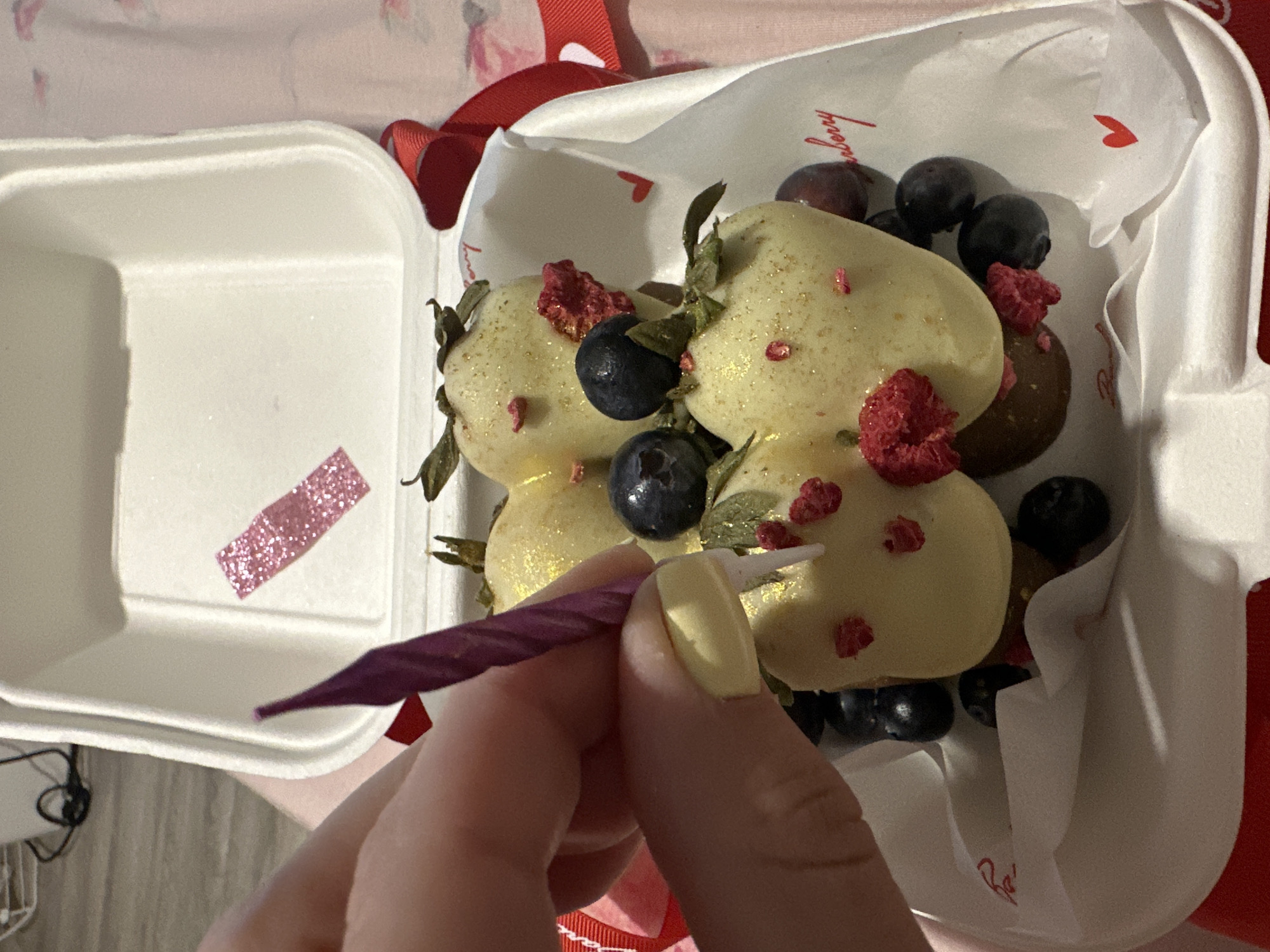 The image depicts a close-up of a white cardboard takeout container filled with white chocolate-covered strawberries and blueberries. A hand with pale skin and light yellow nail polish is holding a purple birthday candle, inserting it into one of the strawberries. The strawberries are dusted with what appears to be gold glitter and adorned with small red pieces, possibly dried raspberries. Red ribbons are visible around the container. The background shows a light-colored surface and a blurry pink fabric. The scene appears to be indoors, likely a domestic setting, and there is no indication of the city of Perm, Russia.