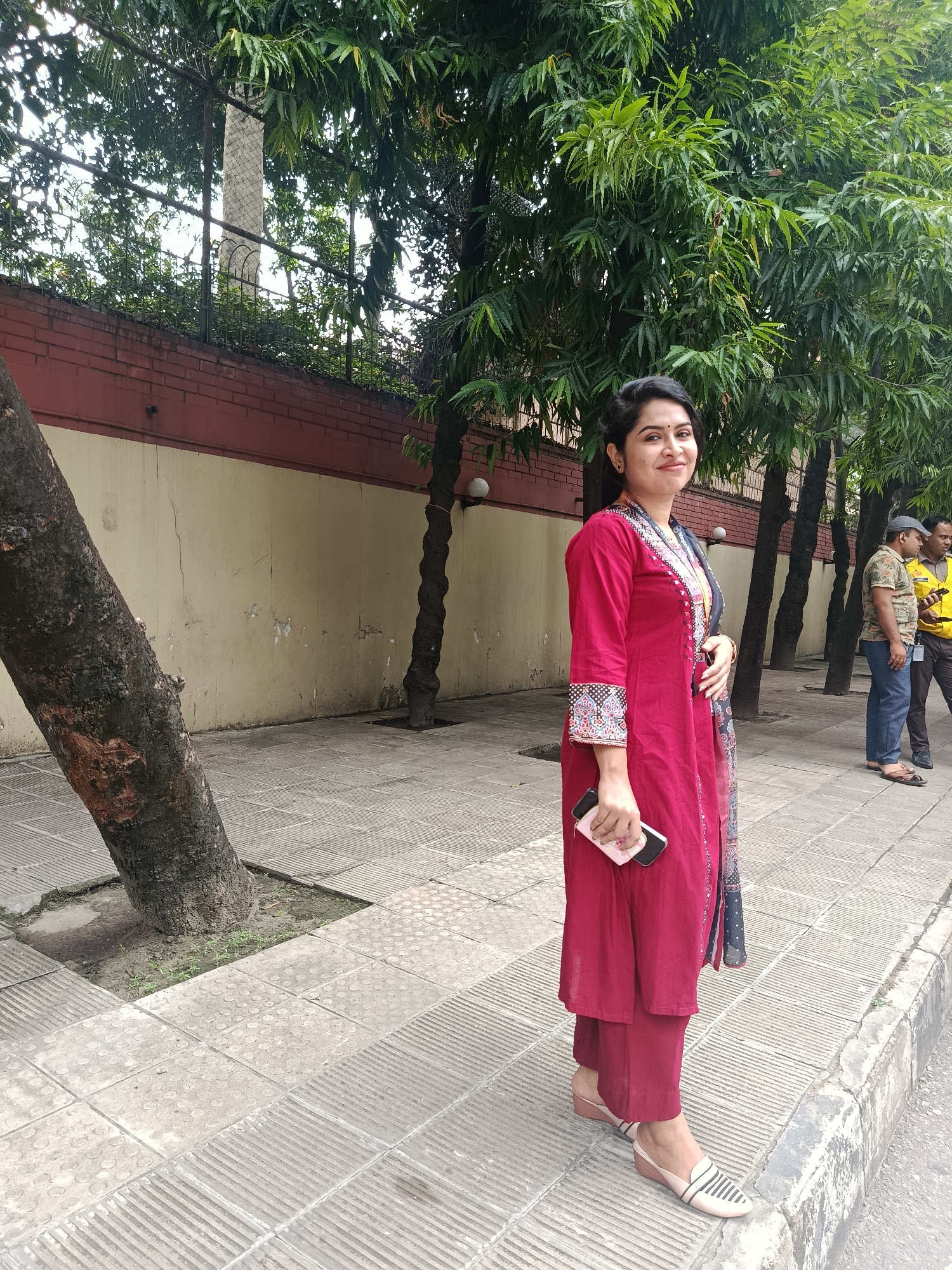 A woman stands on a sidewalk adjacent to a brick and concrete wall beneath a row of tall trees. The woman is wearing a red salwar kameez and holding a smartphone in her right hand. Two men walk behind her on the sidewalk. The scene suggests an urban environment, specifically the city of Dhaka, Bangladesh. The sidewalk consists of gray concrete tiles with textured surfaces. Trees cast shadows across the sidewalk.