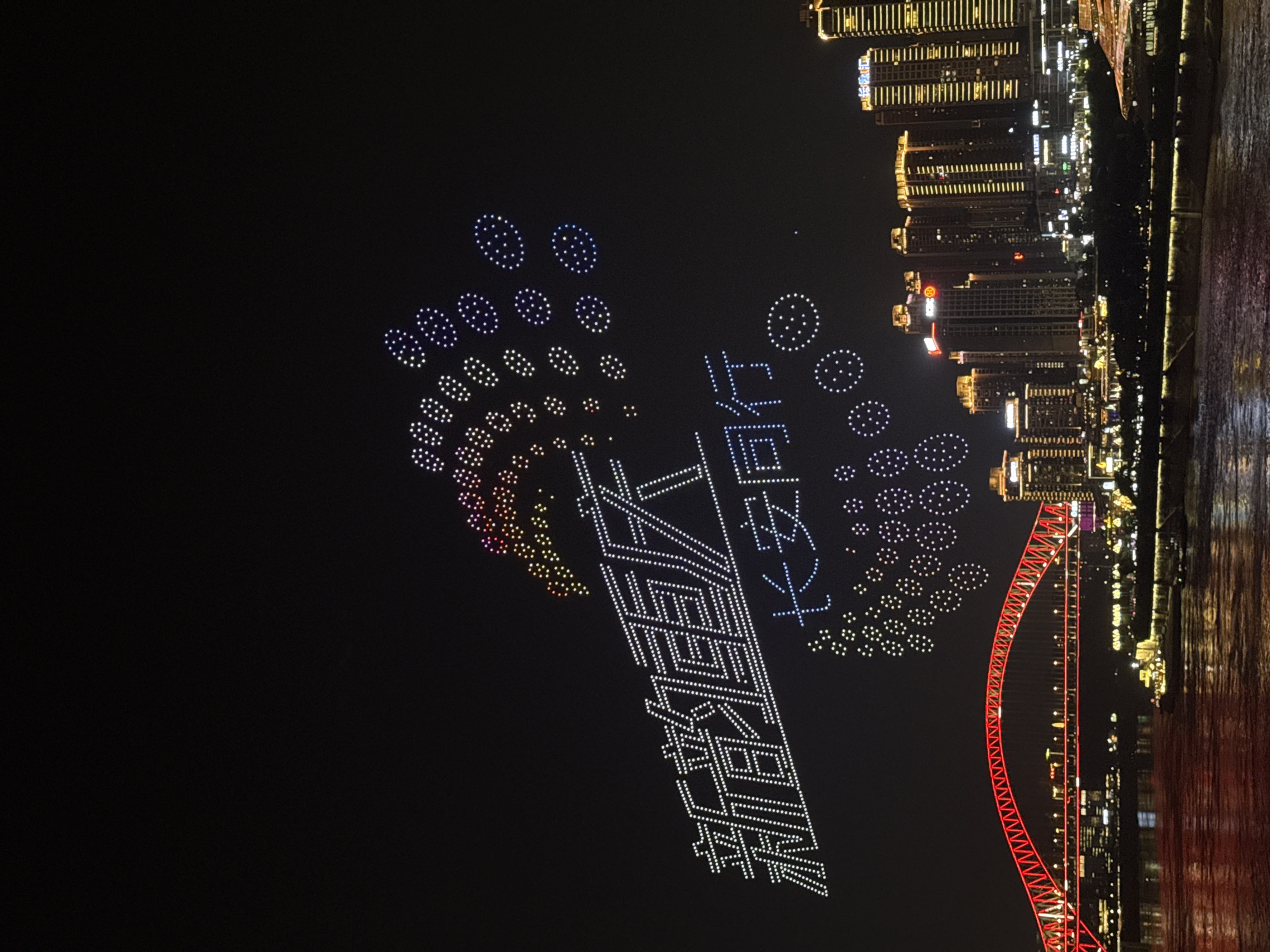 In Yuzhong District, China, a nighttime aerial drone display illuminates the sky above a cityscape and a river. The drone formations form Chinese characters and circular patterns in white and colorful lights. Below the drones, a city skyline is visible with illuminated buildings and a red, arched bridge spanning a body of water. A boat is present on the water. The scene is characterized by the contrast between the dark night sky and the vibrant lights of the drones, city, and bridge.