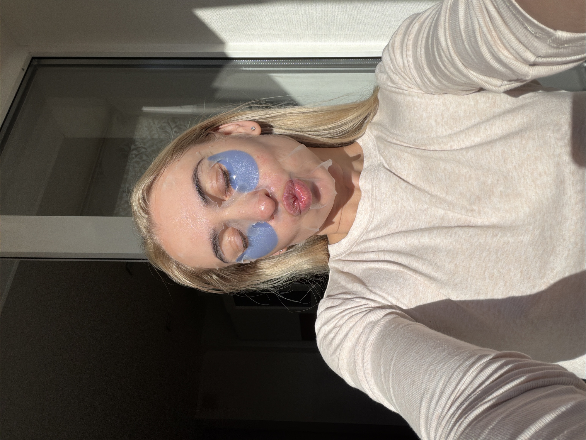A young Caucasian woman with blonde hair and closed eyes is taking a selfie. She is wearing a light-colored, long-sleeved shirt and has blue under-eye patches on. Her face is wet and appears to be covered in a sheet mask. She is puckering her lips. The background shows a dimly lit interior with a window that has strong sunlight casting shadows. This scene is not specific to Chaikovsky, Russia, and depicts a common at-home beauty routine.